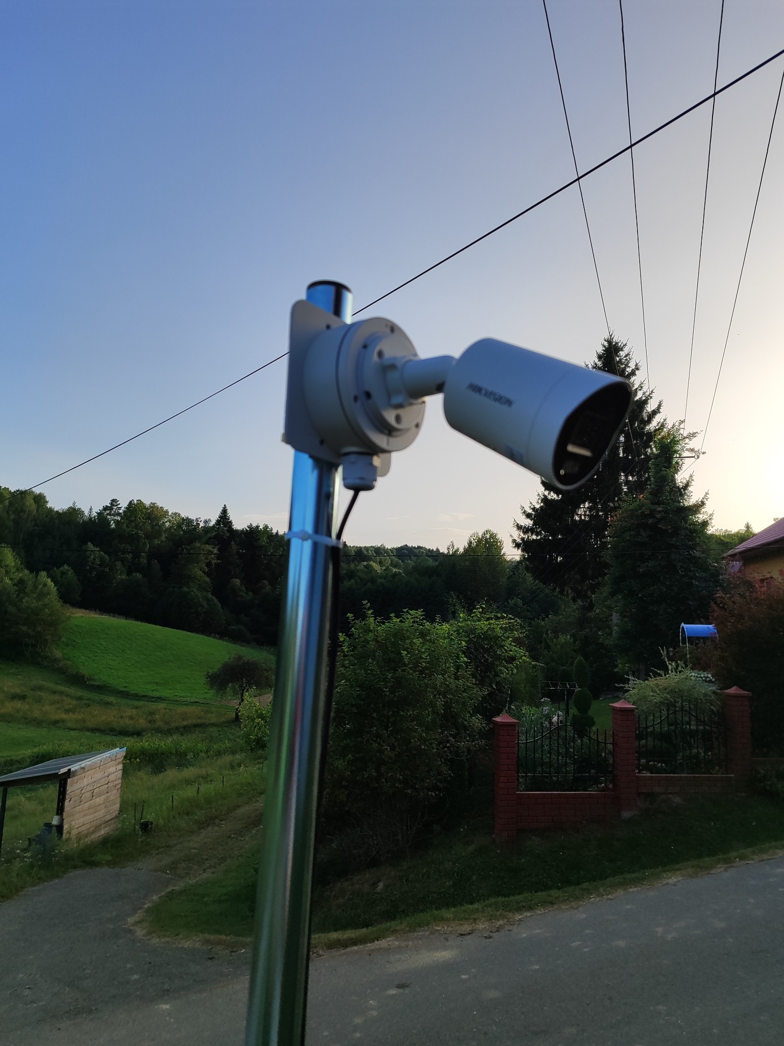 Kamera monitoringu Hikvision na metalowym słupku, zamontowana na zewnątrz z widokiem na zielony krajobraz i dom. Widoczne przewody zasilające.