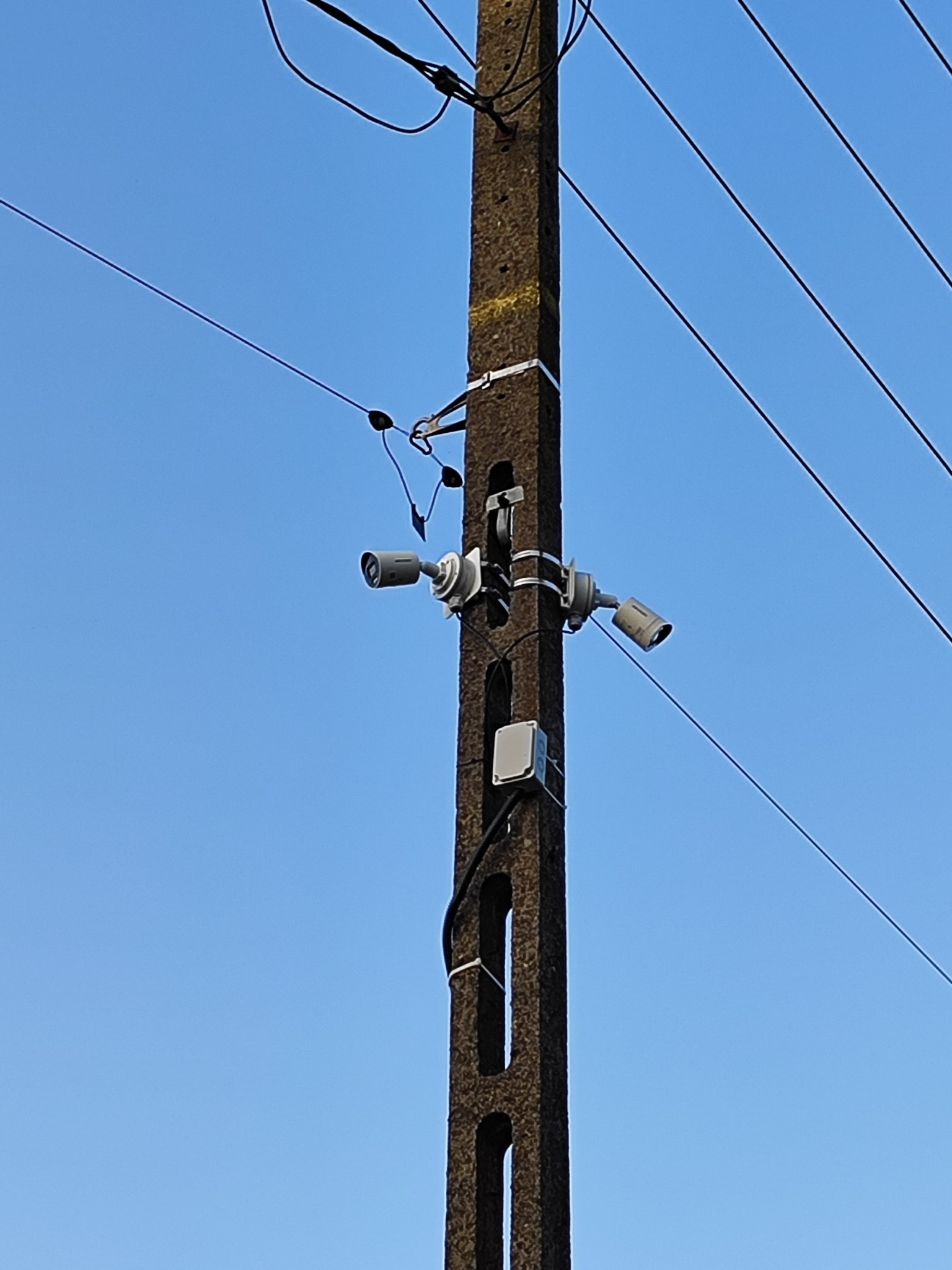 Dwie kamery monitoringu zamontowane na betonowym słupie energetycznym, widoczne przewody i niebieskie tło nieba. Kamery skierowane w przeciwnych kierunkach.