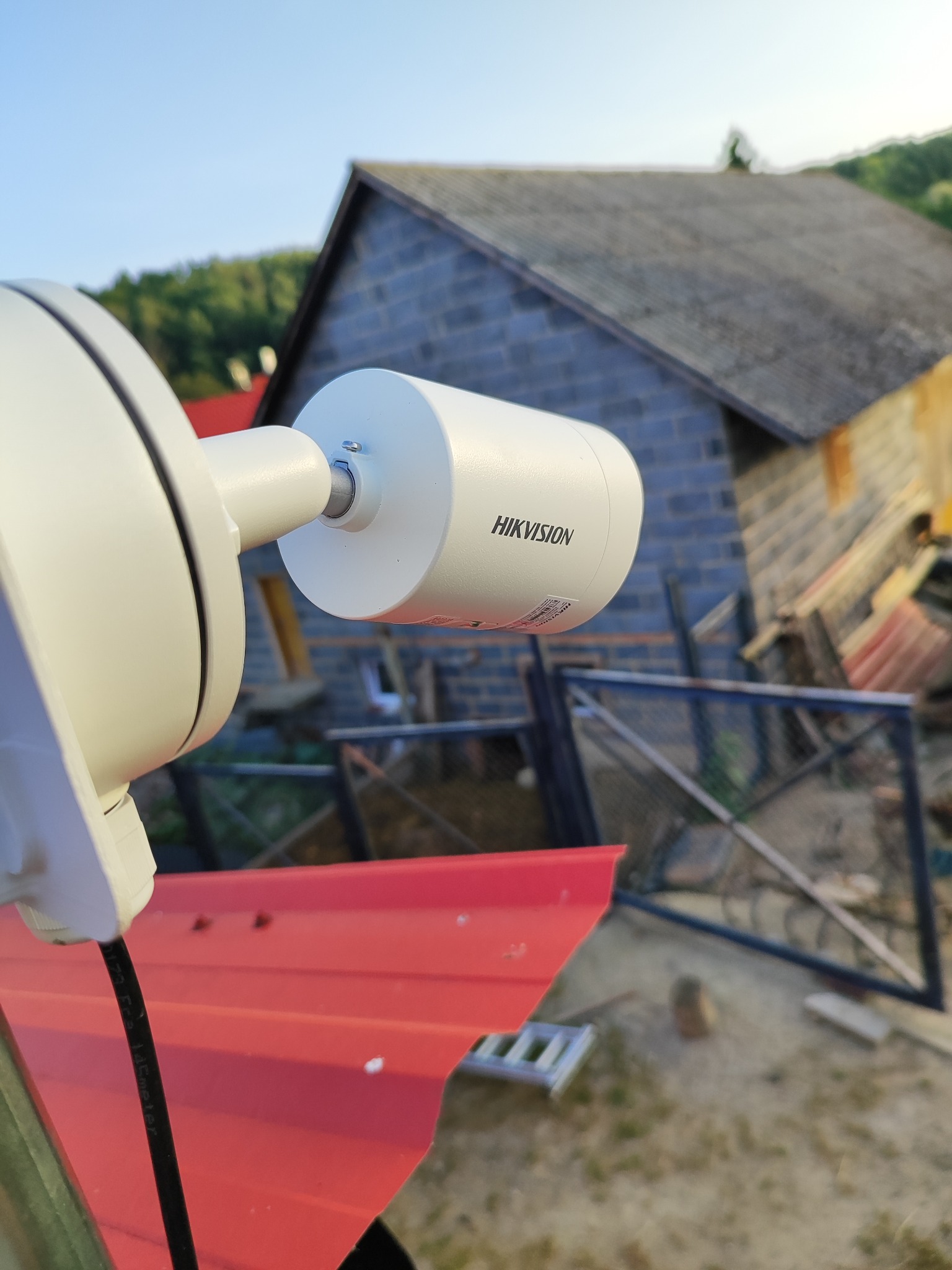 Kamera monitoringu Hikvision zamontowana na czerwonym dachu, z widokiem na budynek w tle. Ujęcie z dołu, podkreślające instalację systemu zabezpieczeń.