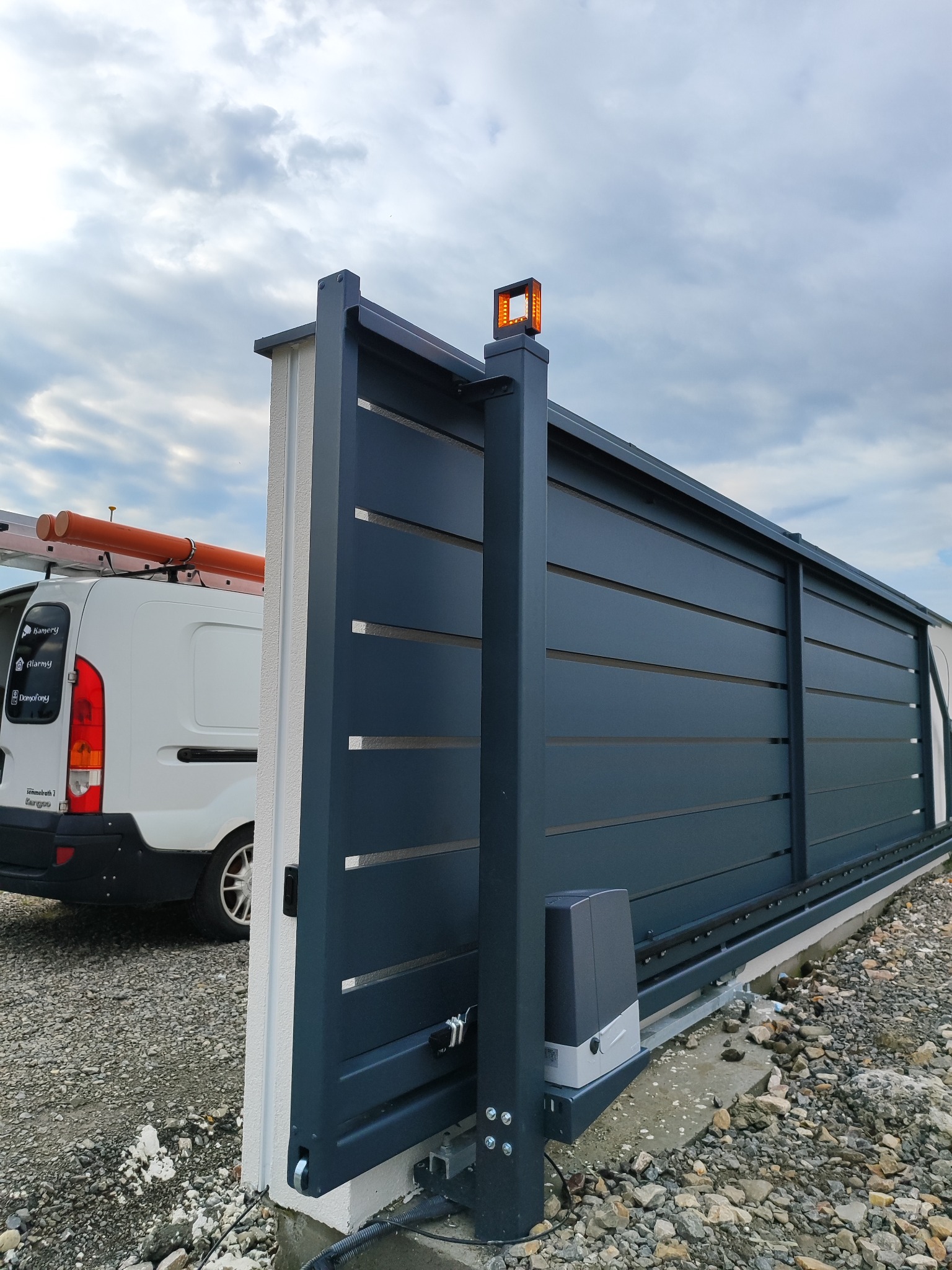 Automatyczna brama przesuwna w kolorze grafitowym, z widocznym napędem i lampą ostrzegawczą. W tle biały bus z drabiną i rurami na dachu, sugerujący montaż lub serwis.