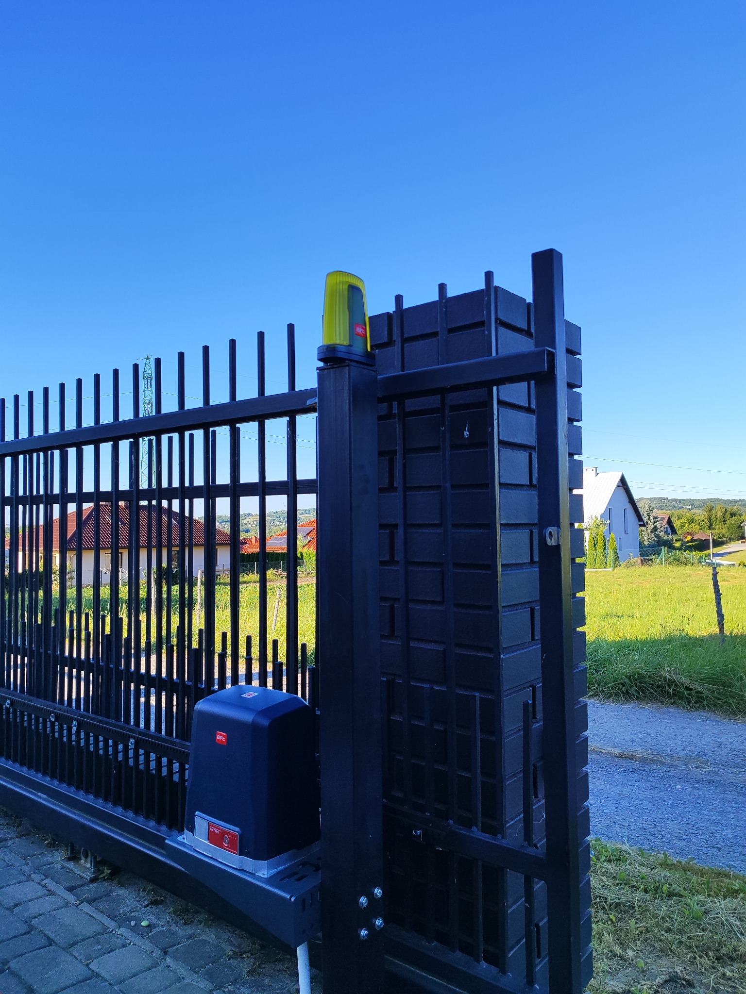Automatyczna brama wjazdowa z napędem BFT, widok z bliska na mechanizm i fragment ogrodzenia. W tle domy i zieleń, błękitne niebo. Nowoczesny design.