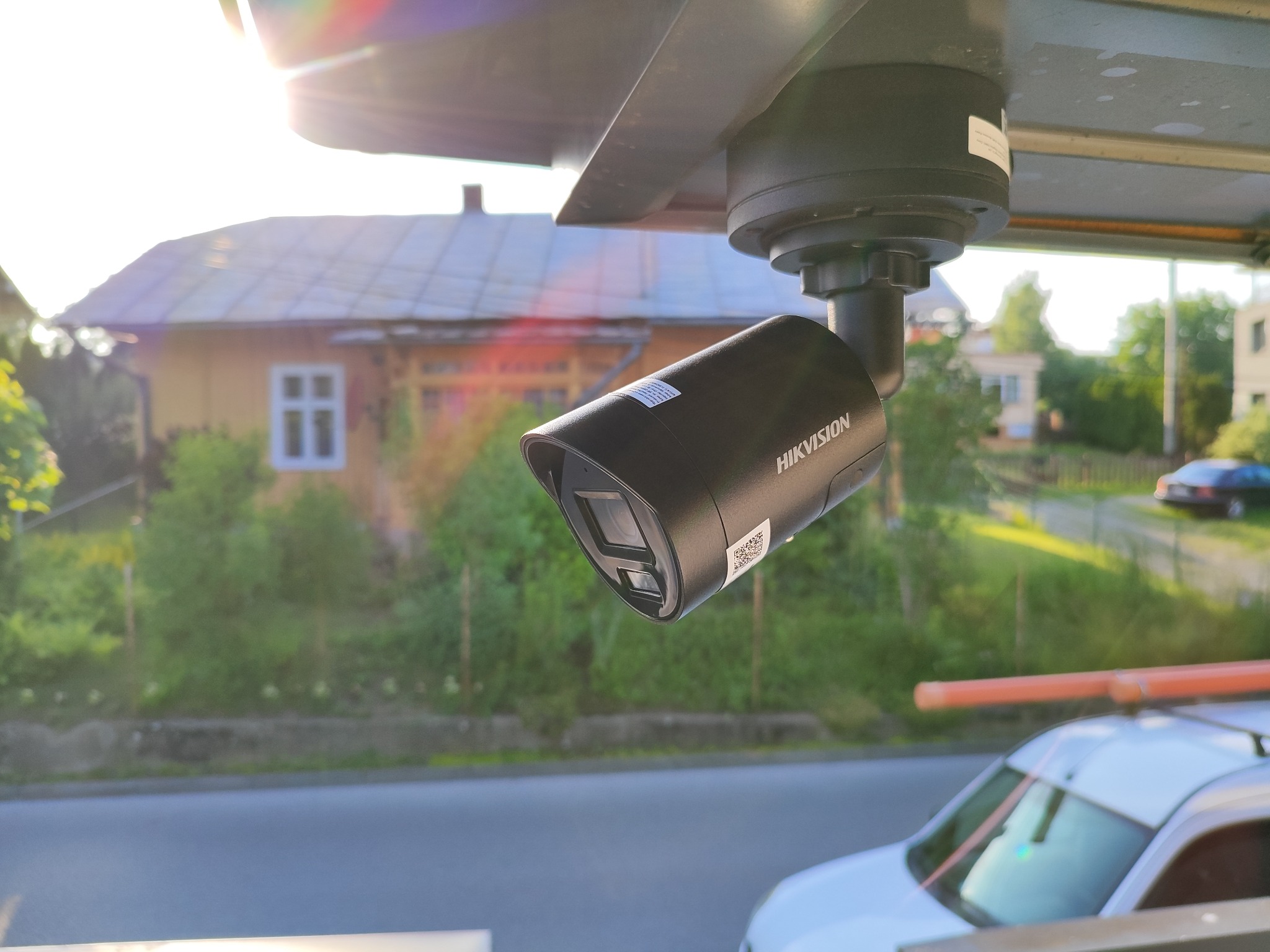 Kamera monitoringu Hikvision zamontowana na zewnątrz budynku, widoczna z dołu, z domem i zielenią w tle. Na dachu samochodu widoczne rury.