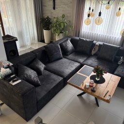 Remigiusz Sokół - Duży, narożny, grafitowy wypoczynek z poduszkami w salonie. Na stoliku bukiet róż. W tle okno z firanami i dekoracyjne lampy.