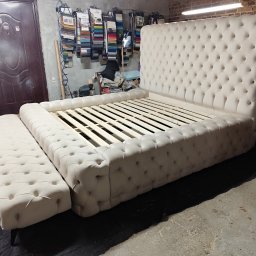 Remigiusz Sokół - Beżowe, tapicerowane łóżko z pikowanym zagłówkiem i podnóżkiem w warsztacie. Widoczne drewniane stelaże i próbki tkanin w tle. Ujęcie z niskiej perspektywy.