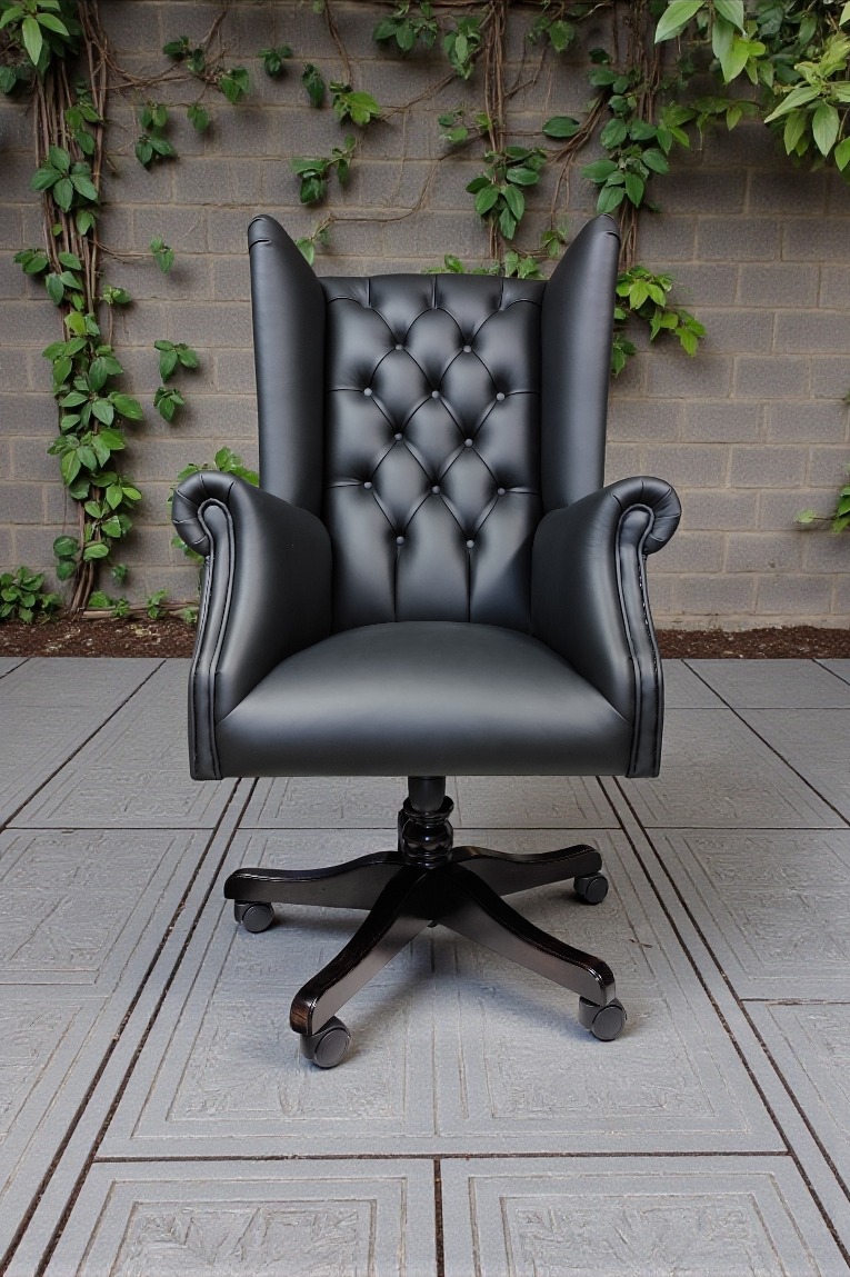 Elegancki, czarny fotel biurowy z pikowanym oparciem, ustawiony na tle ceglanej ściany porośniętej bluszczem. Fotel obrotowy na kółkach, styl Chesterfield.