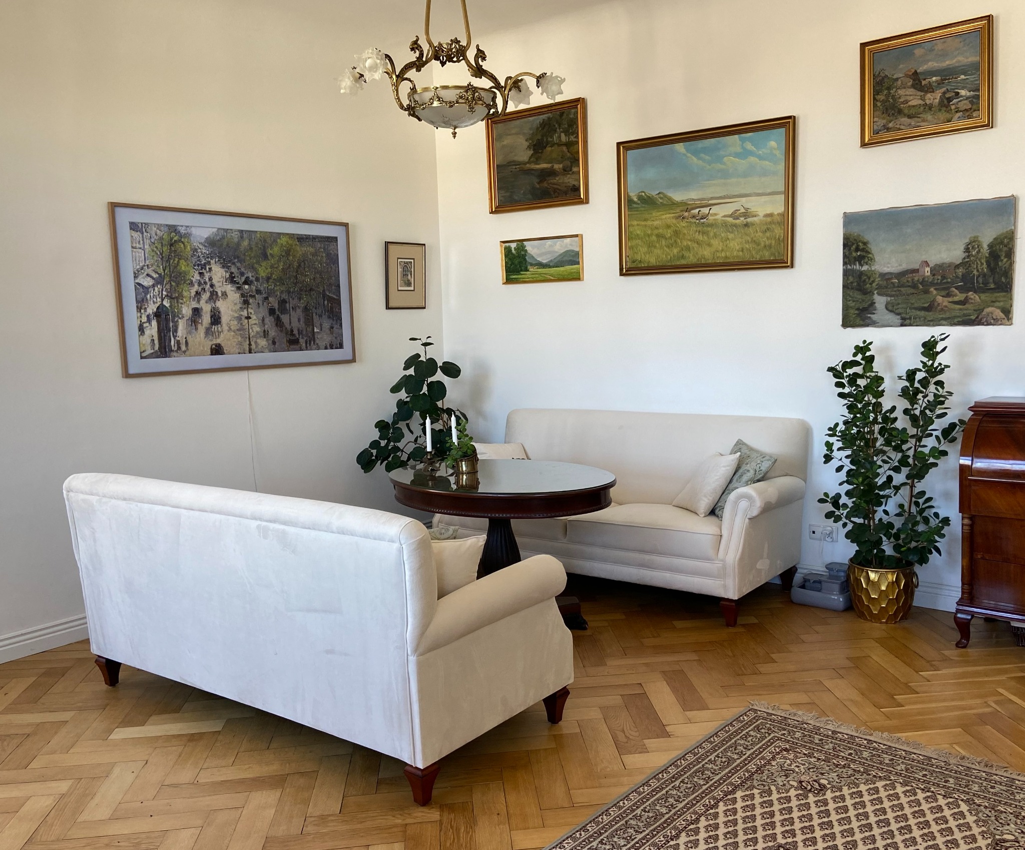 Elegancki salon z dwoma białymi sofami, okrągłym stolikiem i parkietem w jodełkę. Ściany zdobią obrazy w złotych ramach i rośliny doniczkowe.