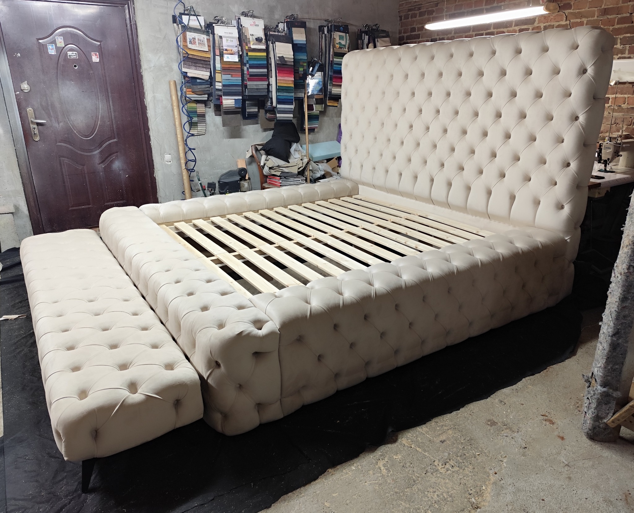 Beżowe, tapicerowane łóżko z pikowanym zagłówkiem i podnóżkiem w warsztacie. Widoczne drewniane stelaże i próbki tkanin w tle. Ujęcie z niskiej perspektywy.