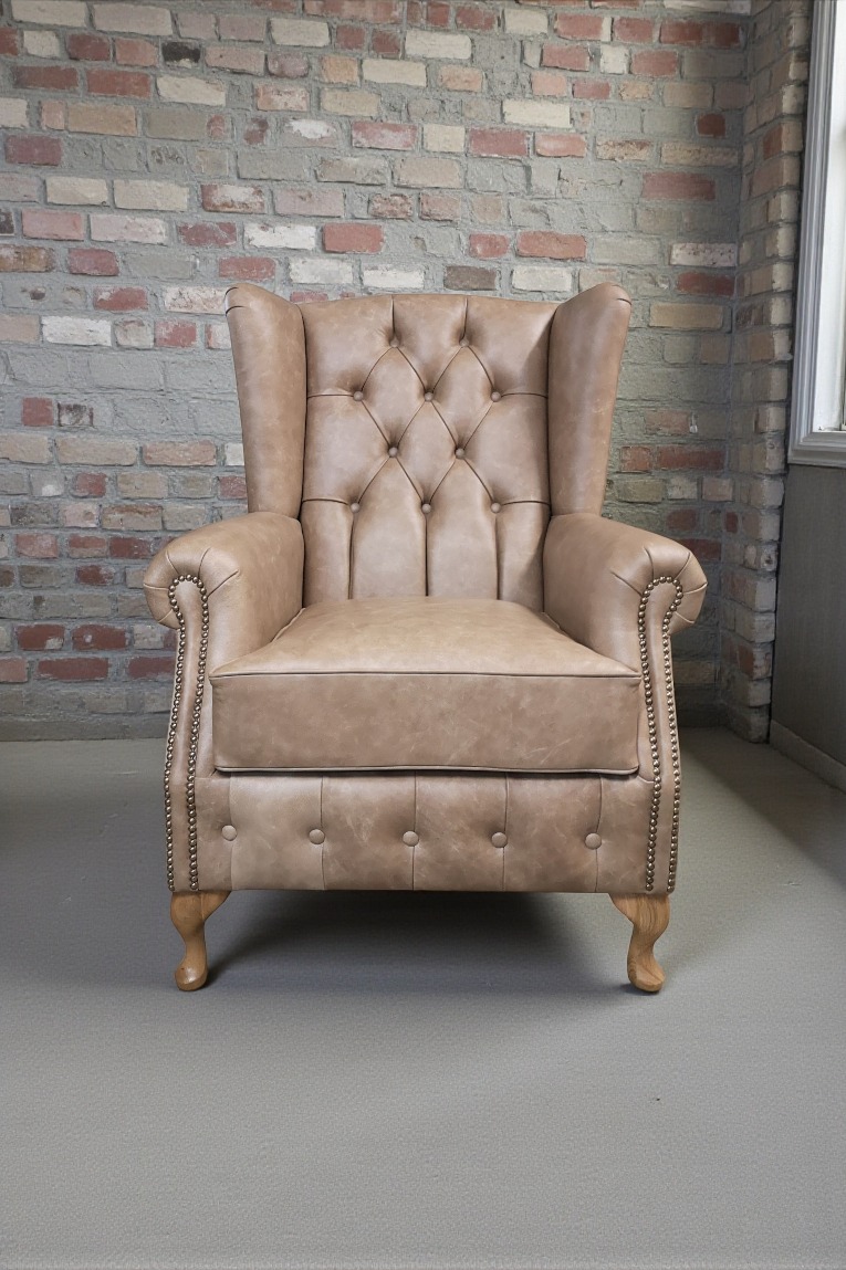 Beżowy fotel typu uszak z pikowanym oparciem i ozdobnymi nitami, ustawiony na tle ceglanej ściany. Widoczne drewniane nóżki i subtelne cieniowanie skóry.