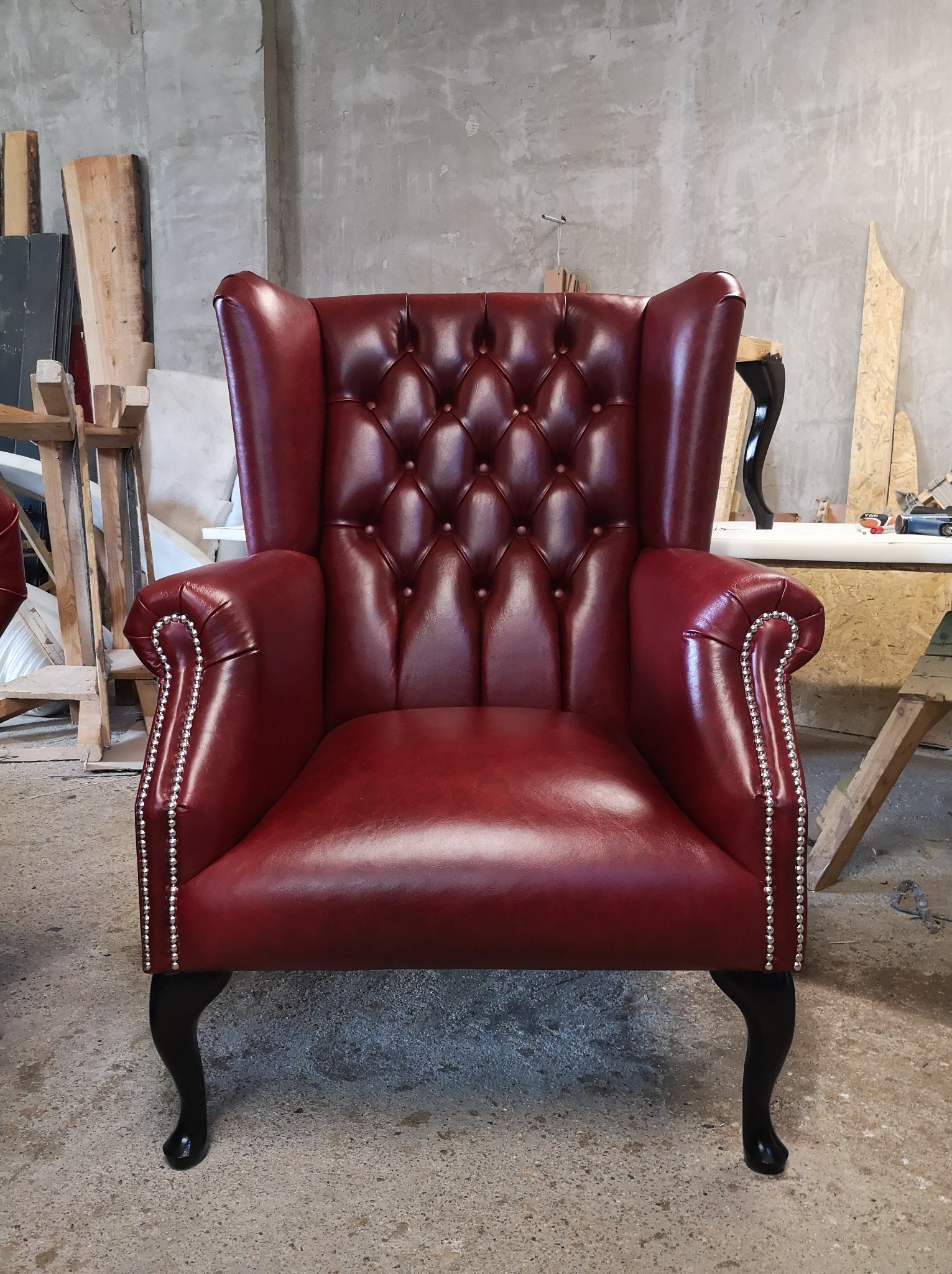 Burgundowy fotel typu uszak z pikowaniem i srebrnymi nitami, stojący w warsztacie na tle surowego betonu. Widoczne drewniane elementy i narzędzia.