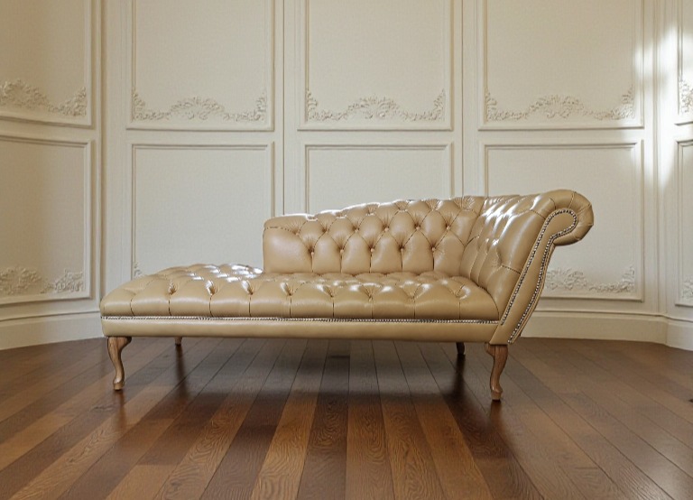 Beżowy, pikowany szezlong z drewnianymi nóżkami na parkiecie, na tle ściany z dekoracyjnymi panelami. Klasyczny styl, elegancki design.