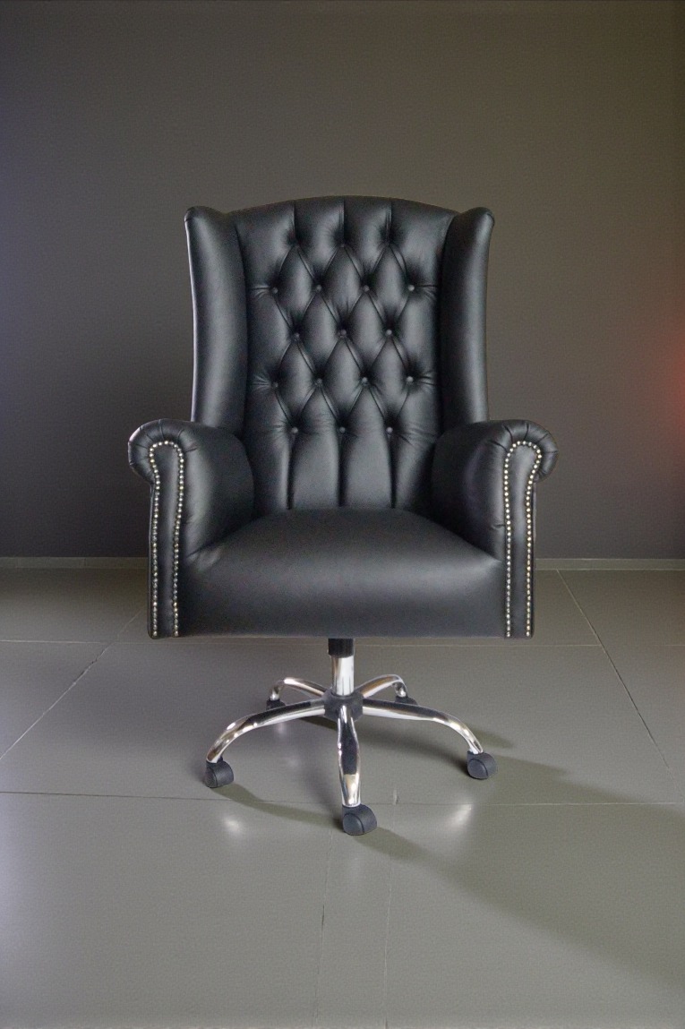 Czarne, skórzane krzesło biurowe w stylu Chesterfield z chromowaną podstawą na tle szarej ściany i podłogi. Widoczne pikowania i ozdobne nity.