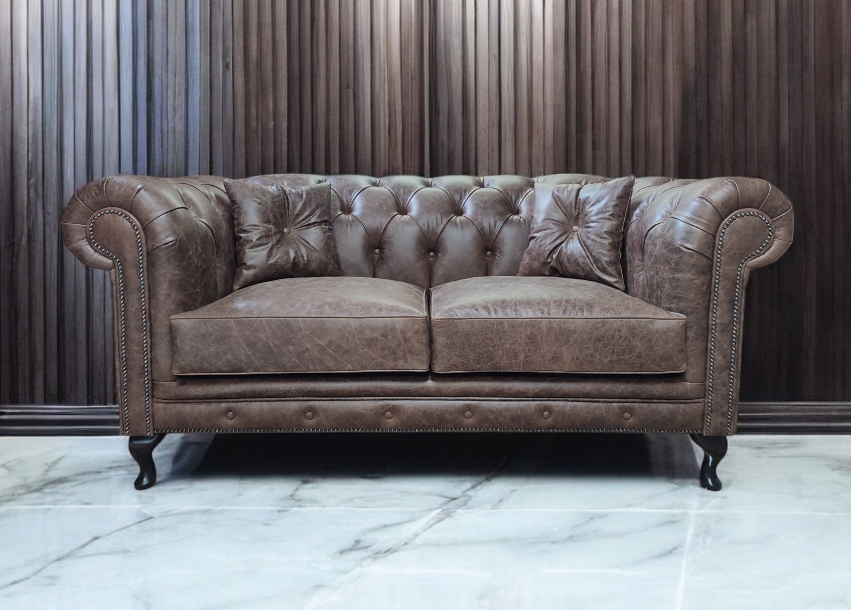 Stylowa, brązowa sofa Chesterfield z dwoma poduszkami na marmurowej podłodze i tle z paneli ściennych. Widoczne detale: nity na podłokietnikach i toczone nóżki.