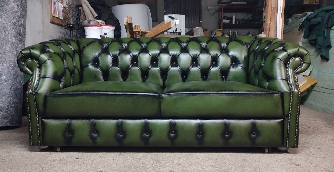 Dwuosobowa sofa Chesterfield w odcieniach zieleni, z charakterystycznym pikowaniem i nitami, na tle warsztatu tapicerskiego. Klasyczny design z nowoczesnym akcentem.