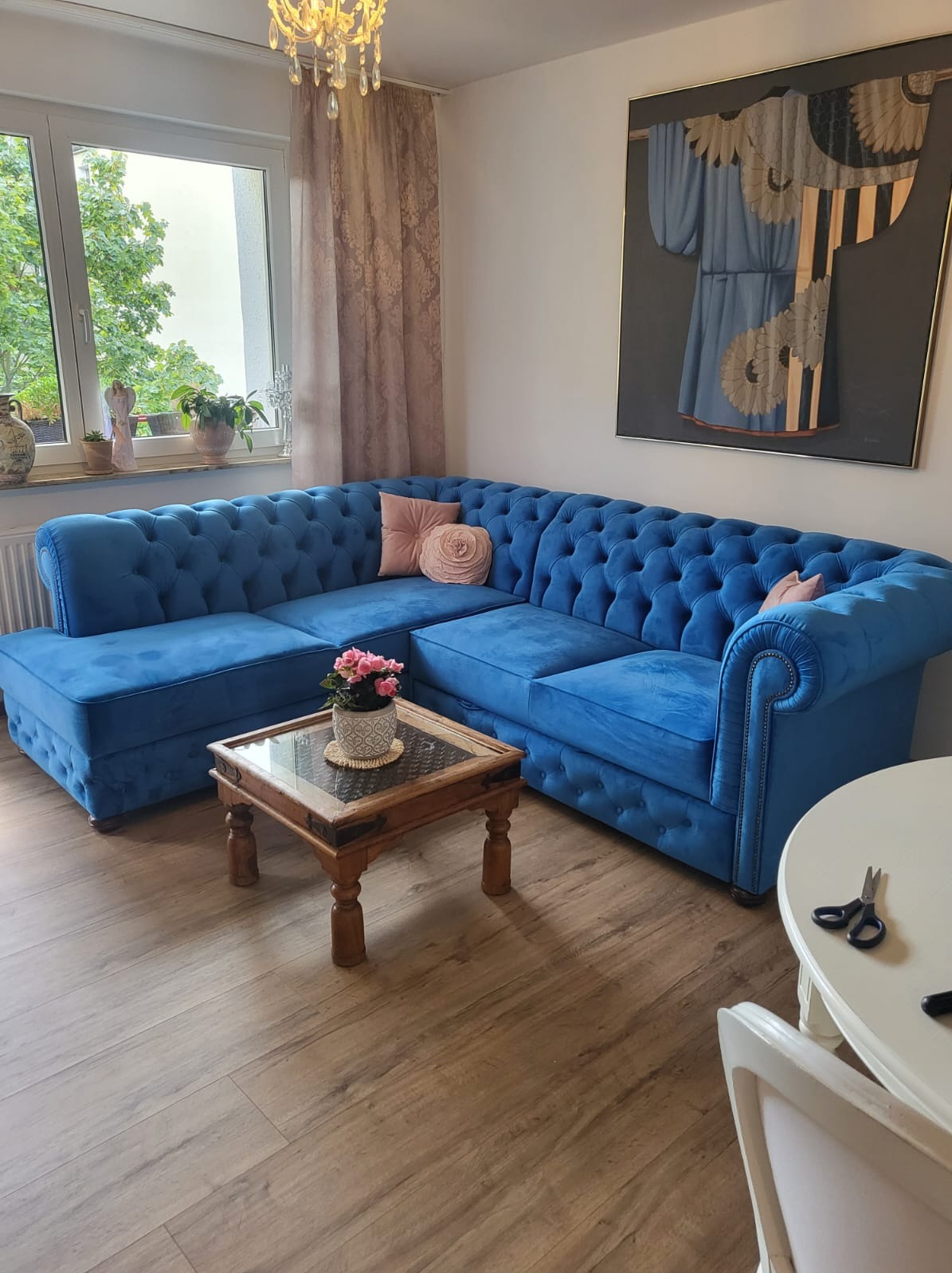 Narożnik Chesterfield w odcieniu błękitu, z dekoracyjnymi guzikami i poduszkami, ustawiony w salonie z drewnianą podłogą i obrazem na ścianie.