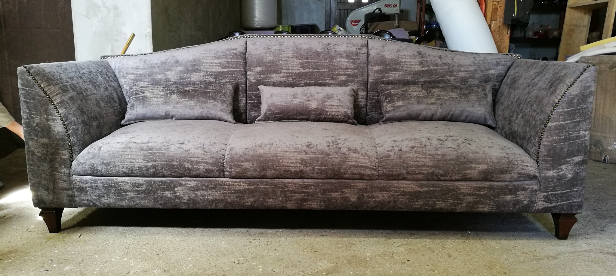 Szary, tapicerowany kanapa z poduszkami, wykończona ozdobnymi gwoździami, na tle warsztatu. Widoczne narzędzia i materiały tapicerskie.