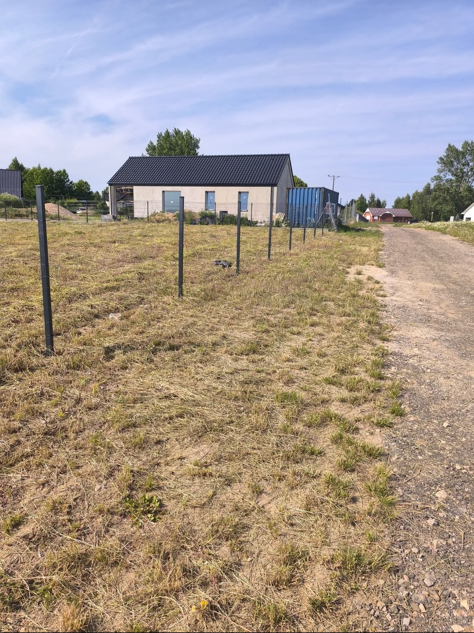 Nowoczesny dom z czarnym dachem w trakcie budowy, otoczony polem z widocznymi słupkami ogrodzeniowymi i drogą gruntową. Błękitne niebo z delikatnymi chmurami w tle.