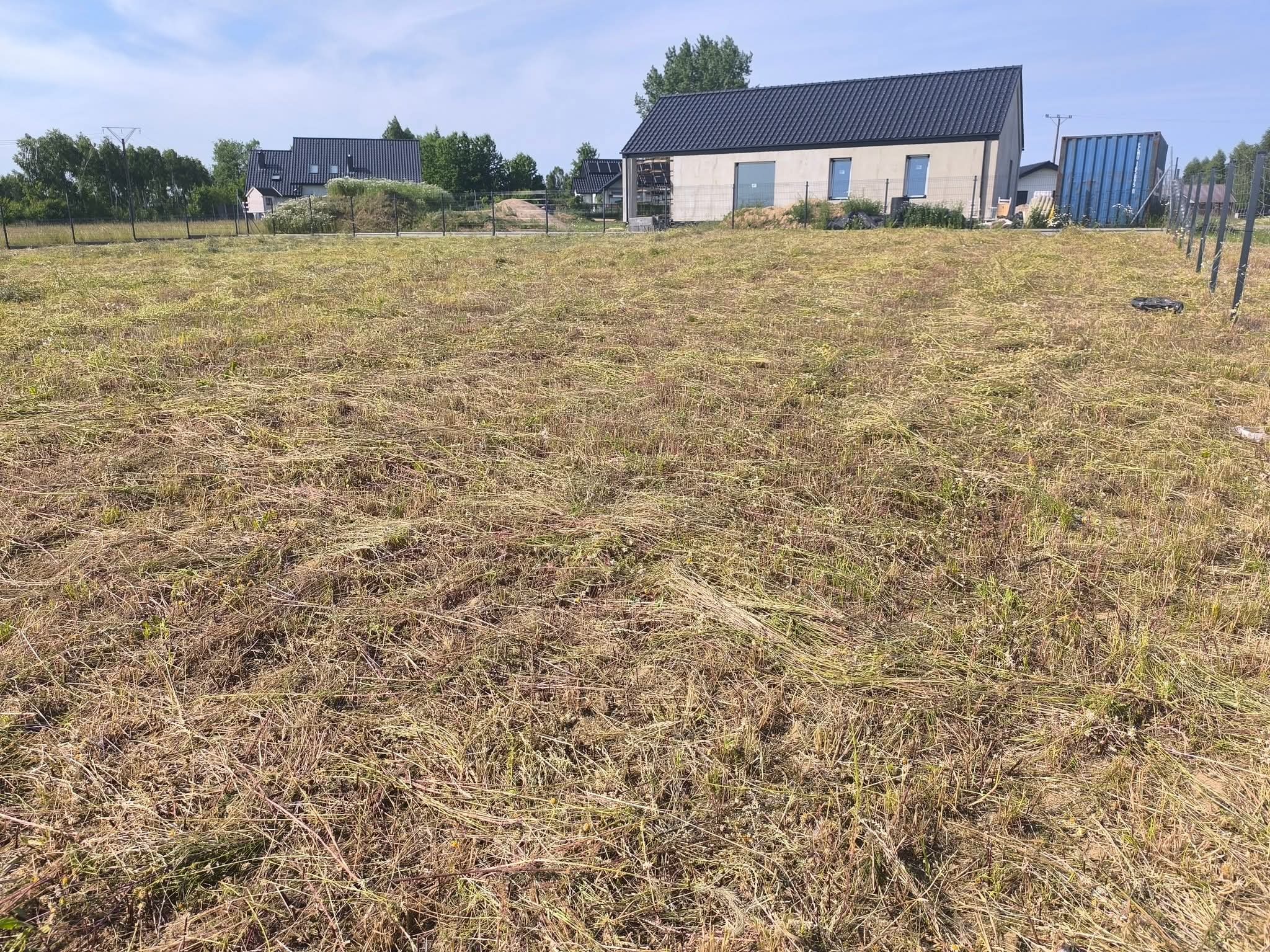 Świeżo skoszone pole z widokiem na nowoczesny dom z ciemnym dachem i szarą elewacją. Na polu leżą pokosy skoszonej trawy. W tle drzewa i słup energetyczny.