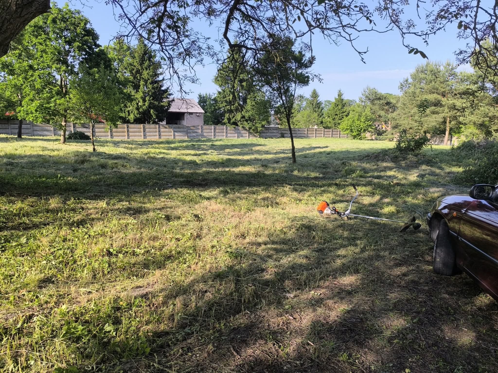 Świeżo skoszone pole z pozostawioną kosiarką spalinową i fragmentem samochodu. W tle widoczny płot i zabudowania. Słoneczny dzień.