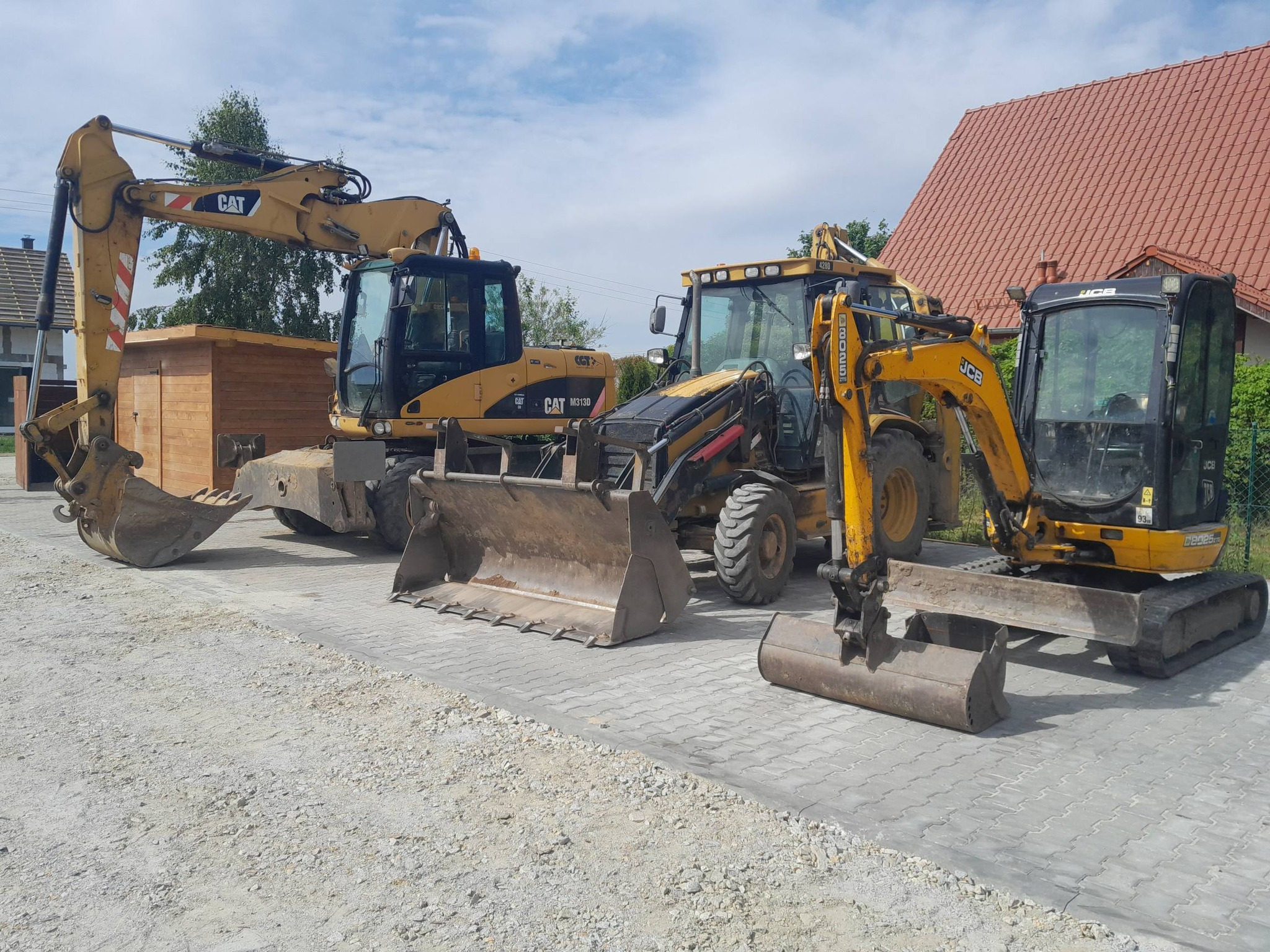 Żółte koparki CAT i JCB stojące na kostce brukowej przed budynkiem z czerwoną dachówką. Sprzęt gotowy do pracy, widoczne łyżki koparek.