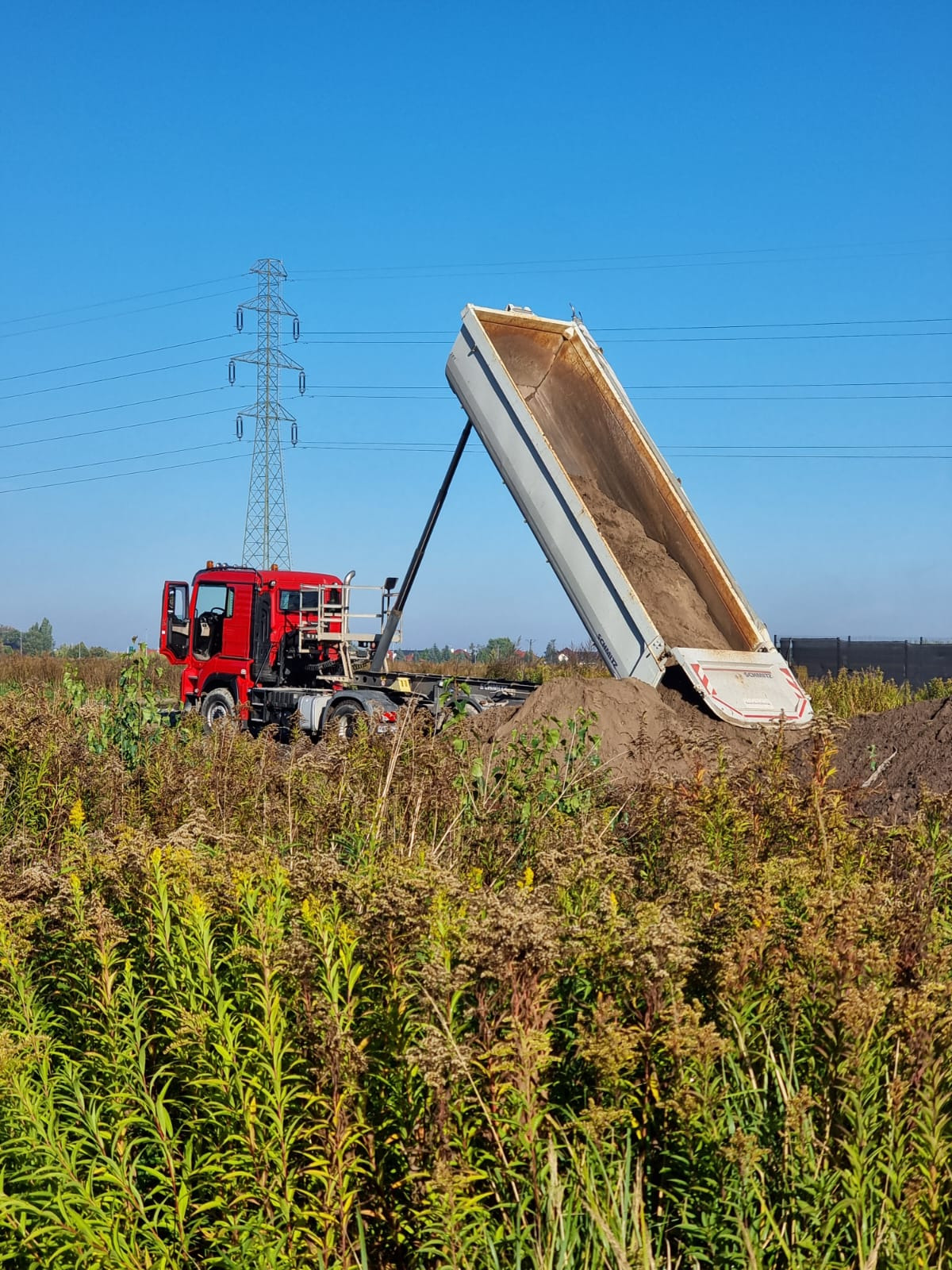 Czerwona wywrotka Schmitz z otwartą skrzynią, wysypująca ziemię na zarośniętym terenie. W tle słup wysokiego napięcia i błękitne niebo.