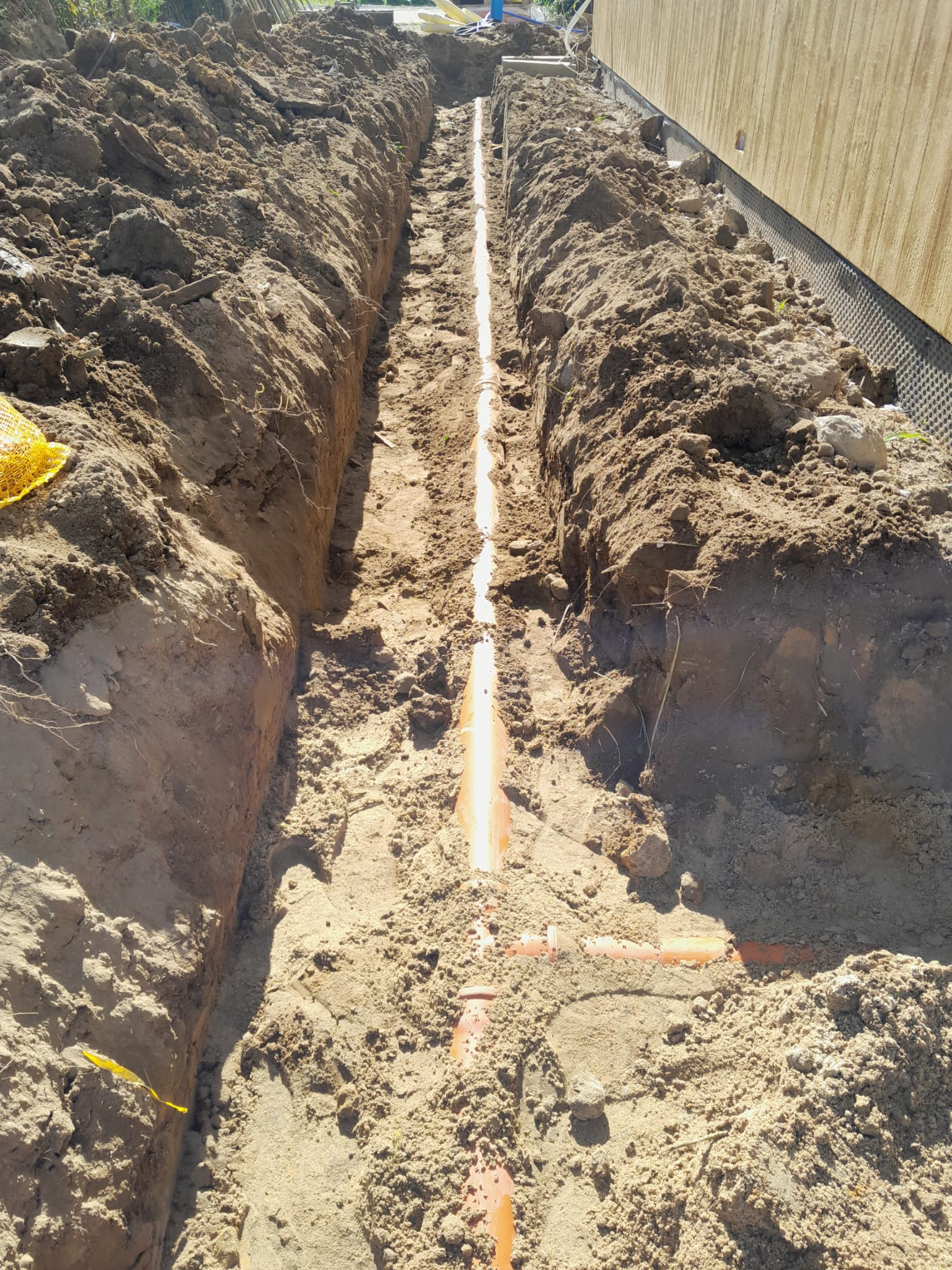 Wąski wykop w ziemi z ułożonymi pomarańczowymi rurami, widok z góry. Ściany wykopu z wyraźną strukturą ziemi, obok budynku z widoczną izolacją fundamentów.