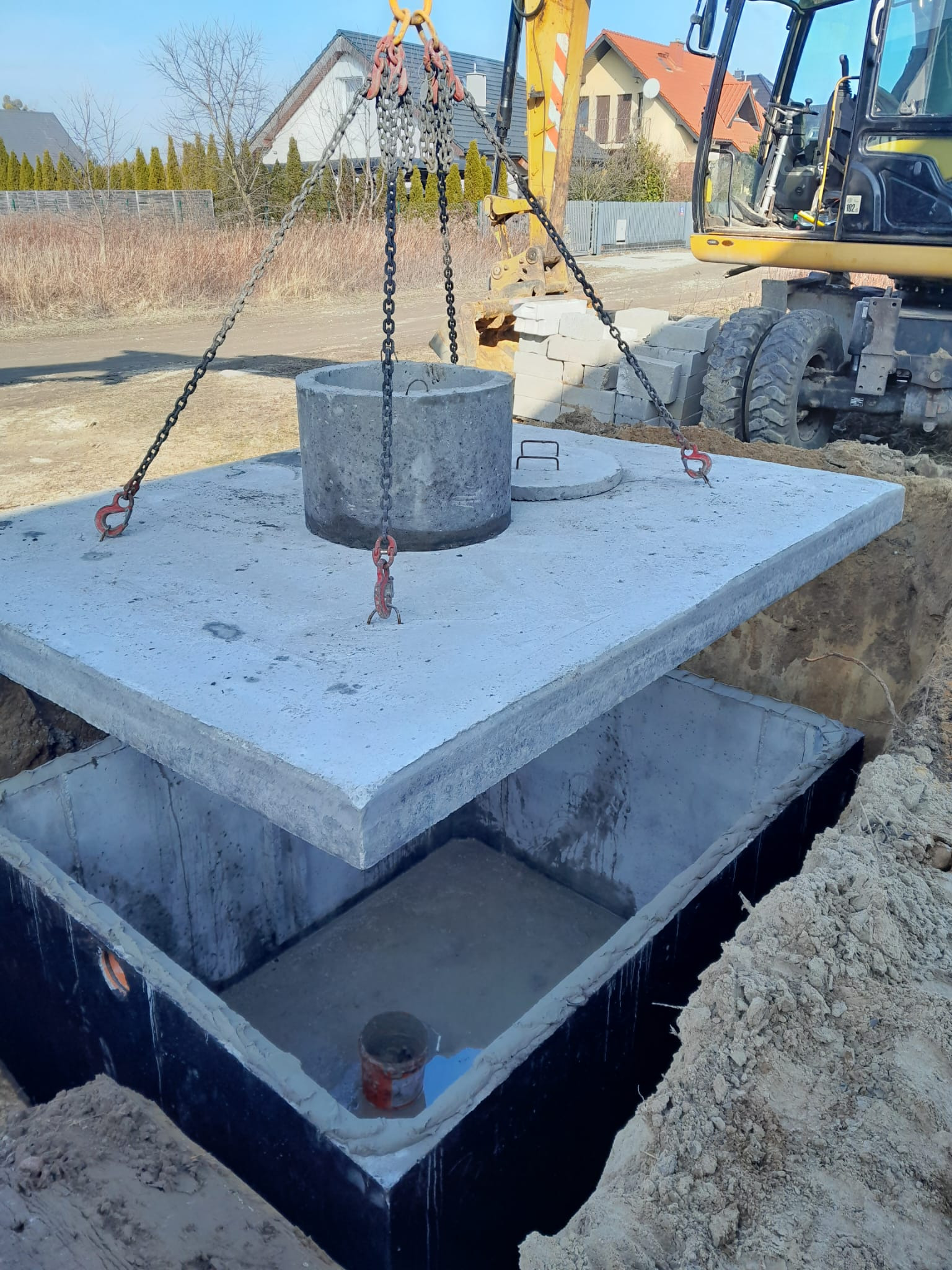 Betonowy zbiornik, prawdopodobnie szambo, w wykopie. Żuraw umieszcza pokrywę. W tle domy i błękitne niebo. Widoczny fragment koparki.