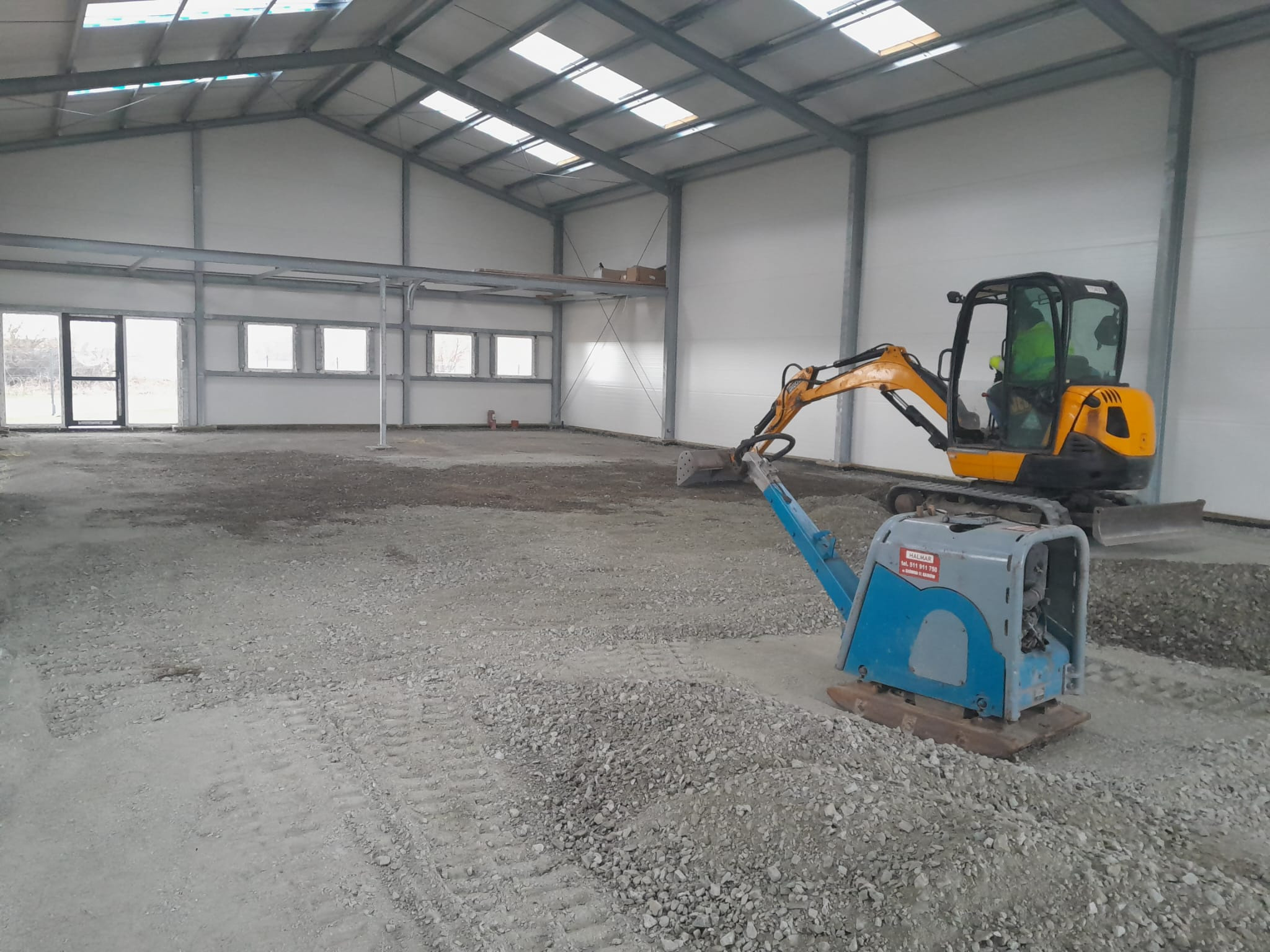 Wnętrze hali z ubitym gruzem, przygotowane pod wylewkę. Widoczny minikoparka i zagęszczarka wibracyjna. Oświetlenie naturalne przez okna dachowe i boczne.