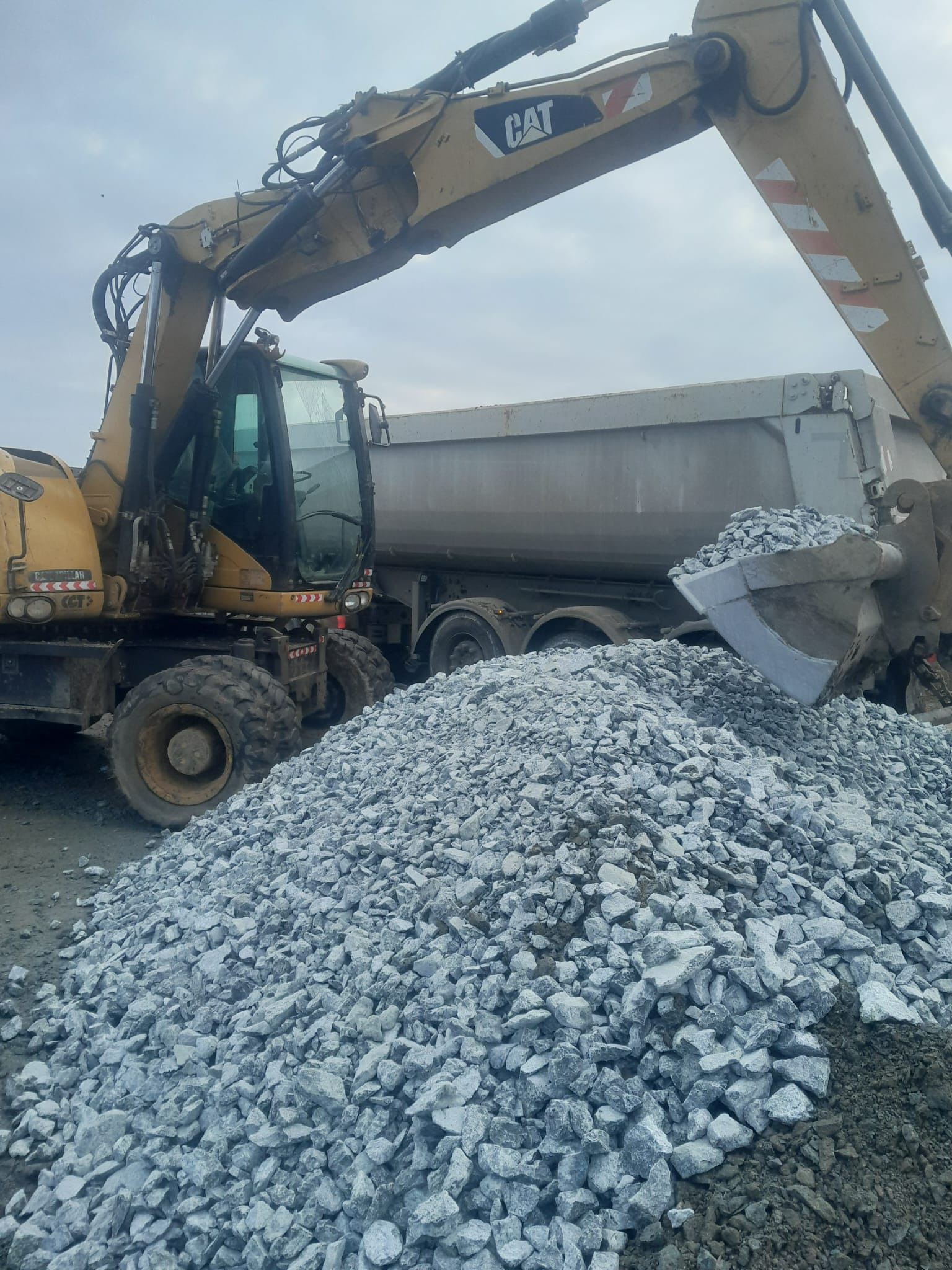 Żółta koparka CAT z łyżką pełną kruszywa, obok wywrotka. Widok z bliska na placu budowy, pochmurne niebo w tle. Kruszywo wysypane na ziemię.