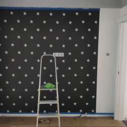 Usługi ogólnobudowlane Dominik kuś - Ściana pomalowana w czarno-białe gwiazdy, zabezpieczona taśmą malarską. Aluminiowa drabina z zieloną szmatką stoi przy ścianie. Widoczny fragment czarnej ściany i białe drzwi.