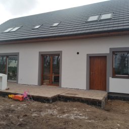 Usługi ogólnobudowlane Dominik kuś - Nowoczesny dom jednorodzinny z szarą stolarką okienną i grafitową dachówką. Widoczne zabawki dziecięce na niezagospodarowanym terenie przed budynkiem.
