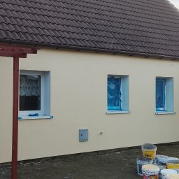 Usługi ogólnobudowlane Dominik kuś - Żółta elewacja domu jednorodzinnego po malowaniu, z widocznymi oknami zabezpieczonymi folią i puszkami farb na trawniku. Otwarta, ciemna brama wejściowa.