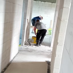 Usługi ogólnobudowlane Dominik kuś - Wnętrze w trakcie budowy: dwóch pracowników w szarych i niebieskich koszulkach, białe ściany z betonu komórkowego, drewniany strop, żółte wiadro i narzędzia.