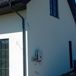 Usługi ogólnobudowlane Dominik kuś - Elewacja domu jednorodzinnego po tynkowaniu, z widocznymi panelami słonecznymi na dachu, oknami z czarnymi ramami i fragmentem ceglanej ściany. Widoczne elementy instalacji elektrycznej.