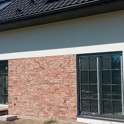 Usługi ogólnobudowlane Dominik kuś - Nowoczesny dom z panelami słonecznymi i fragmentem elewacji wykończonym czerwoną cegłą. Widoczne okna z czarnymi ramami i instalacja elektryczna na zewnątrz.