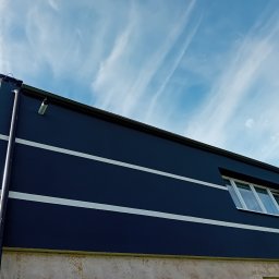 Usługi ogólnobudowlane Dominik kuś - Narożnik budynku z ciemną elewacją, jasnymi pasami i oknem, na tle błękitnego nieba z chmurami. Widoczny fragment rynny i kamery. Ujęcie z dołu.