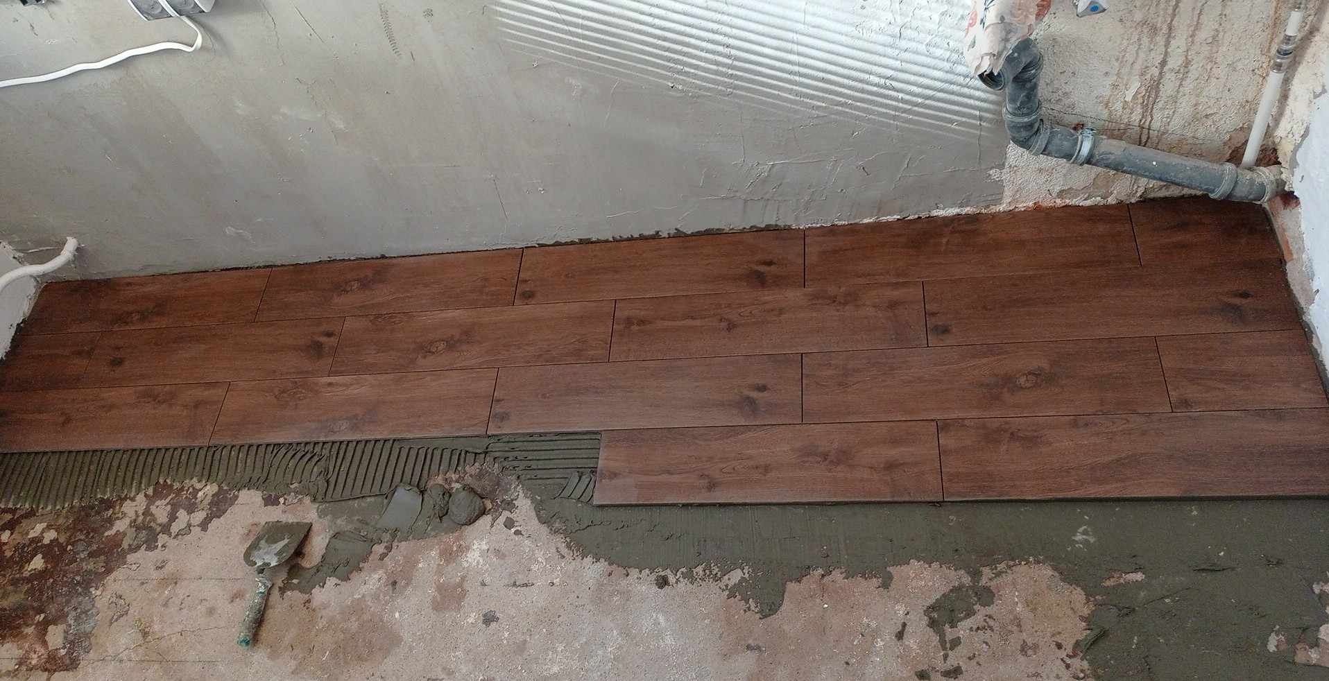 Układanie brązowych paneli podłogowych imitujących drewno, widoczna warstwa kleju i szpachelka. Fragment rury przy ścianie. Prace wykończeniowe w trakcie.