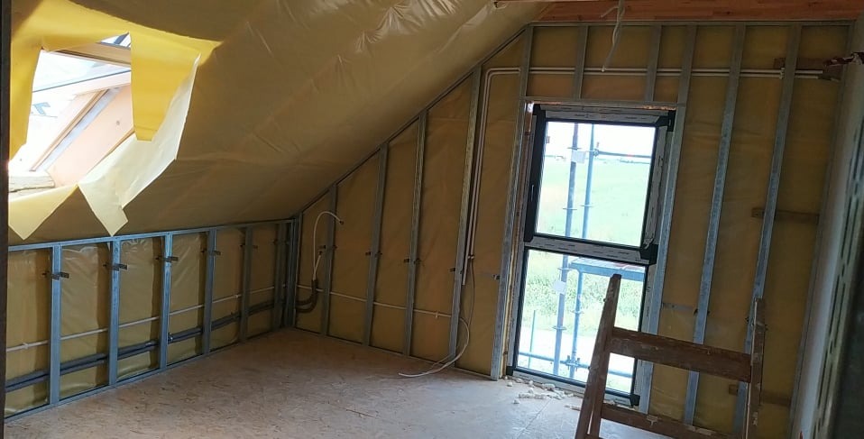 Poddasze w trakcie ocieplania: widoczne metalowe profile, żółta wełna mineralna, okno dachowe i pionowe okno. Częściowo wykończona podłoga z płyt OSB.