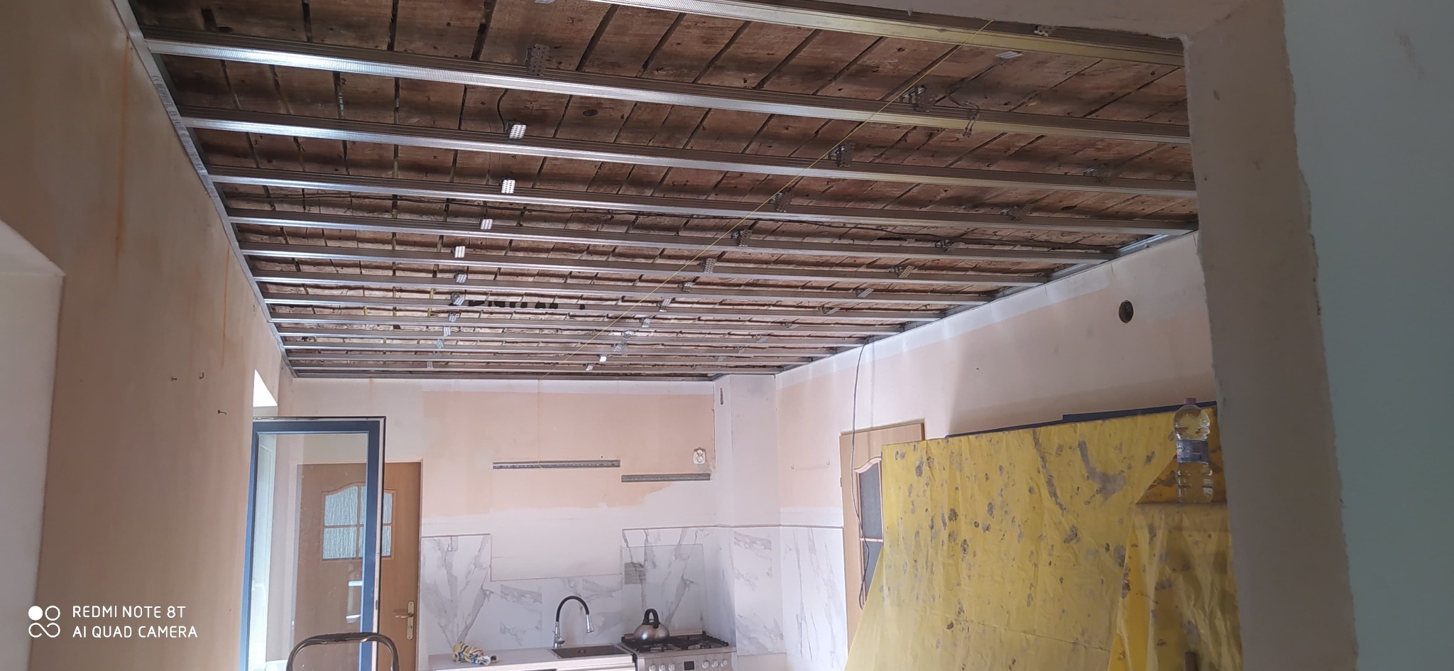 Montaż metalowej konstrukcji sufitu podwieszanego w remontowanym wnętrzu. Widoczne profile, stare drewniane deski i fragment kuchni z białymi płytkami.