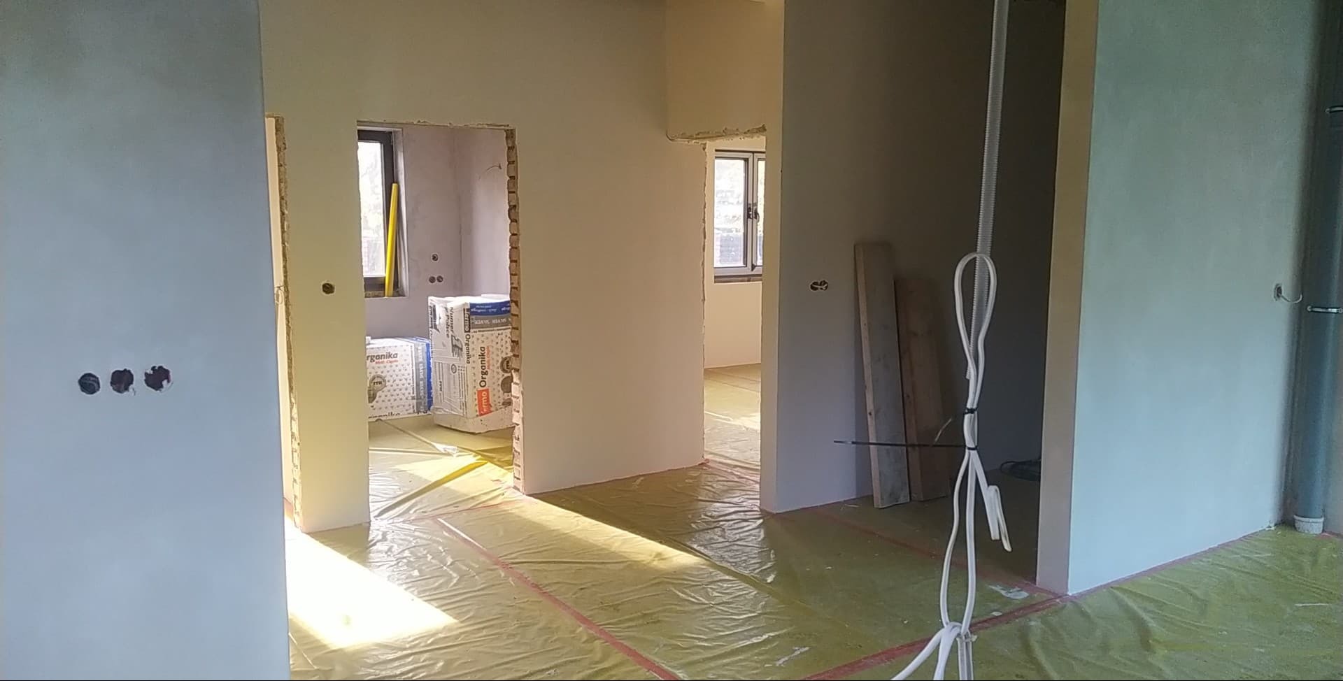 Wnętrze w trakcie prac wykończeniowych: ściany po gładzi, zabezpieczona podłoga folią, widoczne puszki elektryczne i materiały budowlane. Ujęcie z perspektywy wejścia.