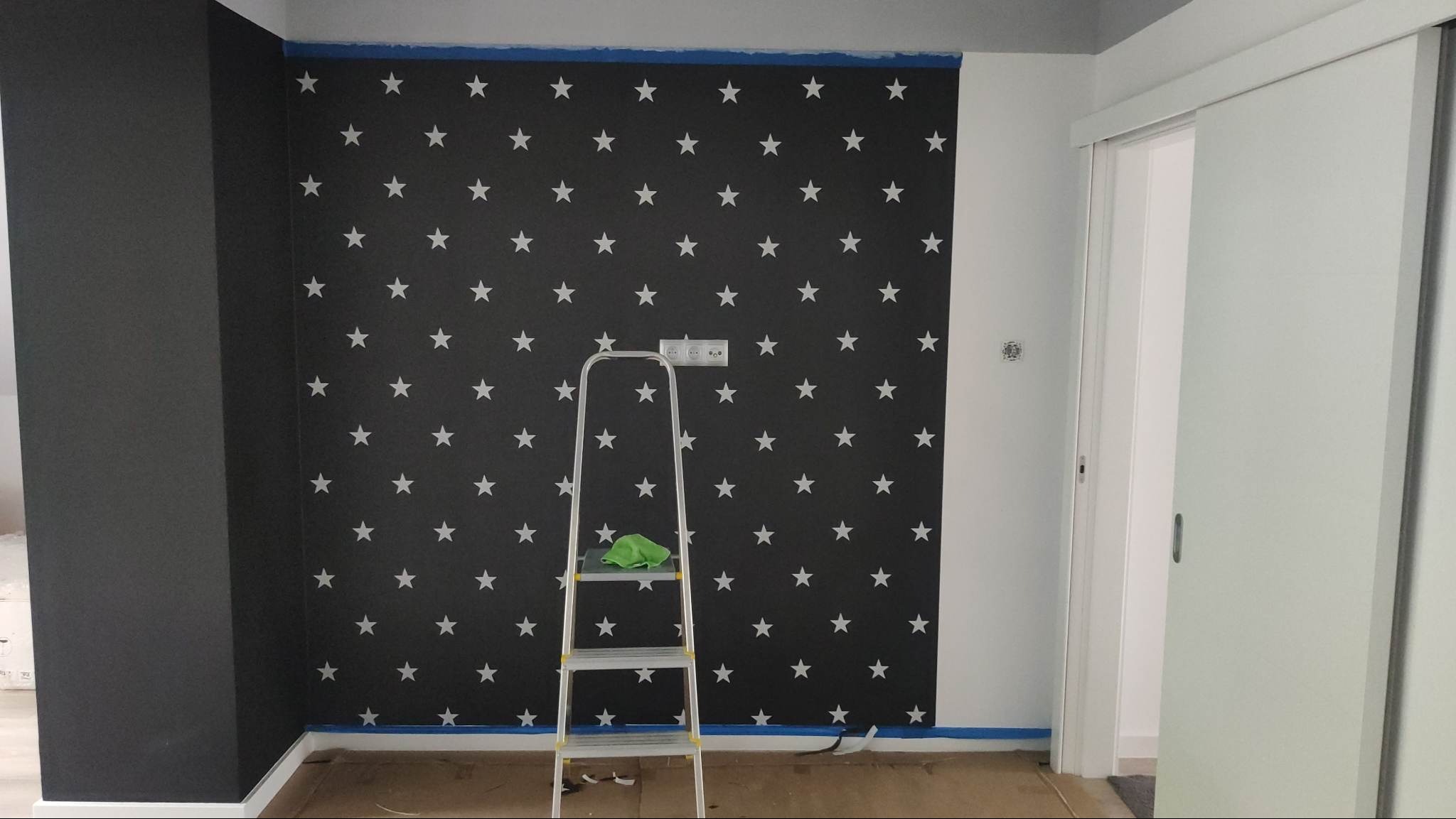 Ściana pomalowana w czarno-białe gwiazdy, zabezpieczona taśmą malarską. Aluminiowa drabina z zieloną szmatką stoi przy ścianie. Widoczny fragment czarnej ściany i białe drzwi.