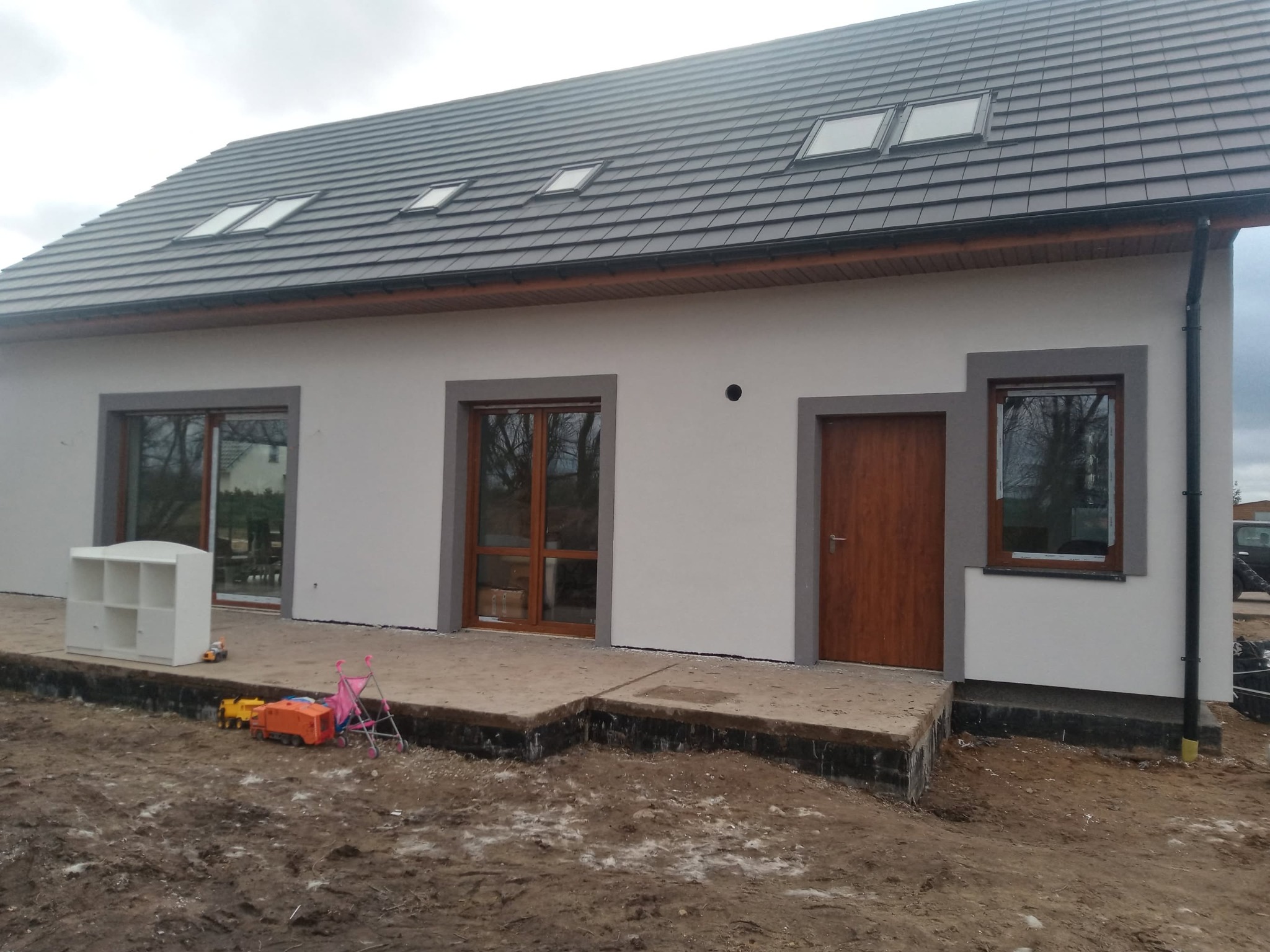 Nowoczesny dom jednorodzinny z szarą stolarką okienną i grafitową dachówką. Widoczne zabawki dziecięce na niezagospodarowanym terenie przed budynkiem.