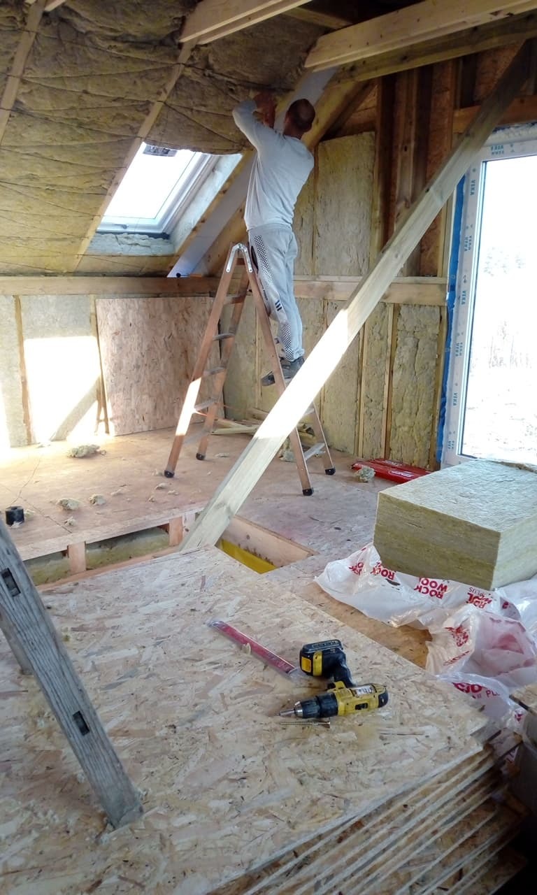 Prace wykończeniowe na poddaszu: ocieplanie wełną mineralną, montaż płyt OSB. Widoczna drabina, narzędzia i okno dachowe. Praca w toku, surowy stan.