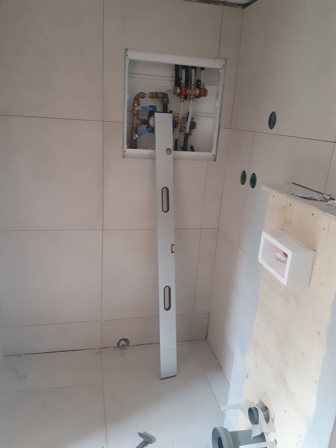 Białe płytki w łazience z instalacją hydrauliczną w ścianie, sprawdzaną poziomicą. Widoczne przyłącza i rury. Częściowo zabudowany stelaż podtynkowy.