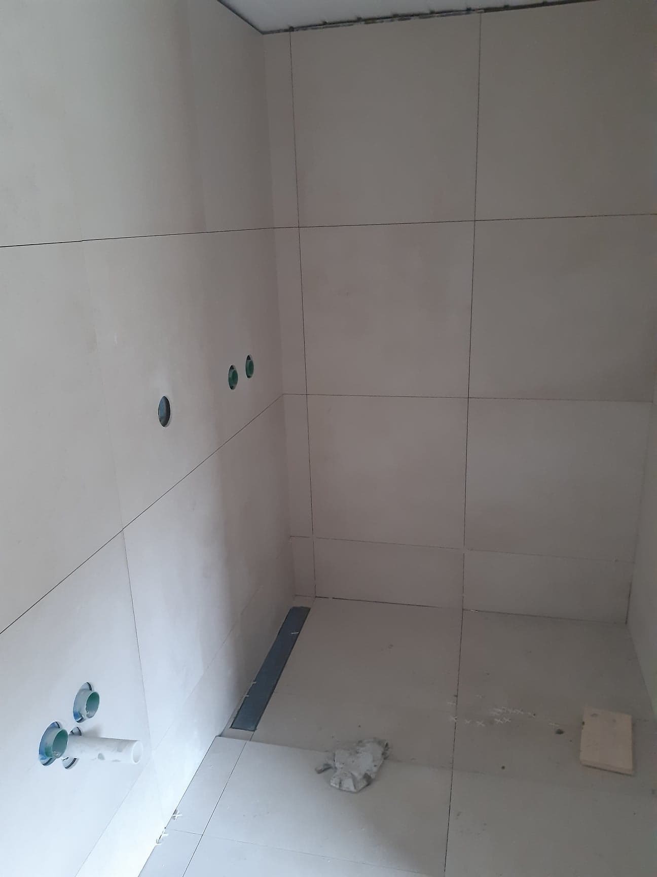 Wnętrze łazienki w trakcie wykańczania: ułożone jasne płytki na ścianach i podłodze, widoczne otwory na instalacje i odpływ liniowy. Minimalistyczny styl.