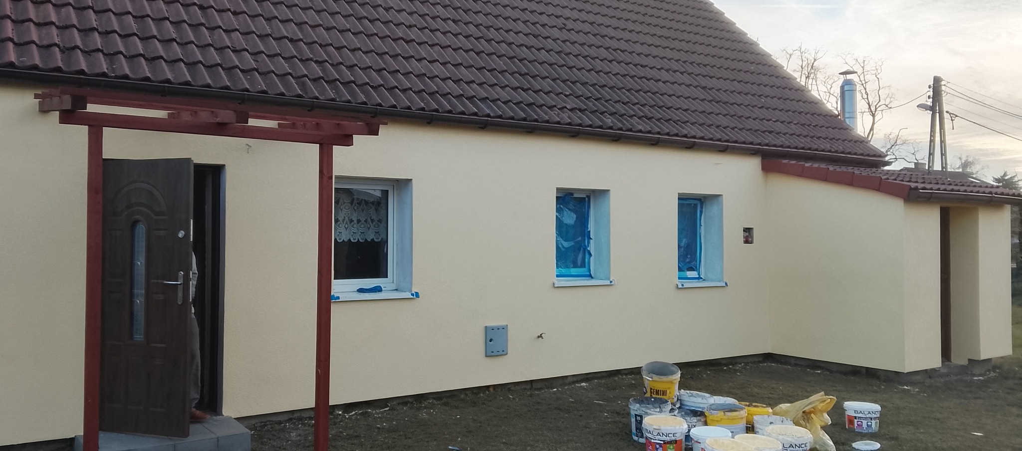 Żółta elewacja domu jednorodzinnego po malowaniu, z widocznymi oknami zabezpieczonymi folią i puszkami farb na trawniku. Otwarta, ciemna brama wejściowa.