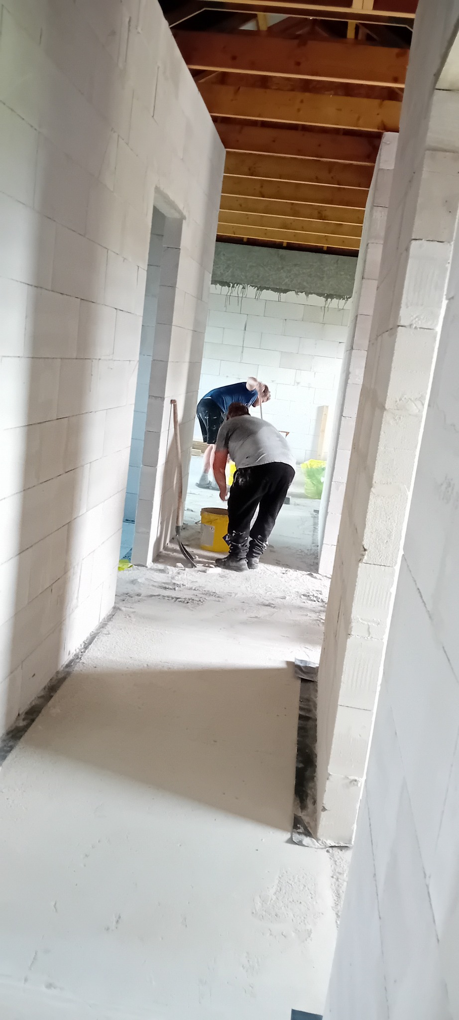 Wnętrze w trakcie budowy: dwóch pracowników w szarych i niebieskich koszulkach, białe ściany z betonu komórkowego, drewniany strop, żółte wiadro i narzędzia.