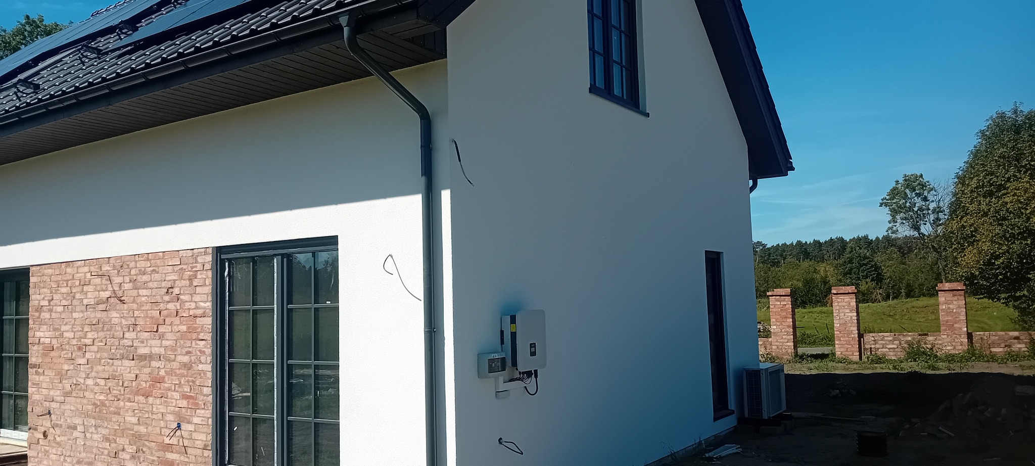 Elewacja domu jednorodzinnego po tynkowaniu, z widocznymi panelami słonecznymi na dachu, oknami z czarnymi ramami i fragmentem ceglanej ściany. Widoczne elementy instalacji elektrycznej.
