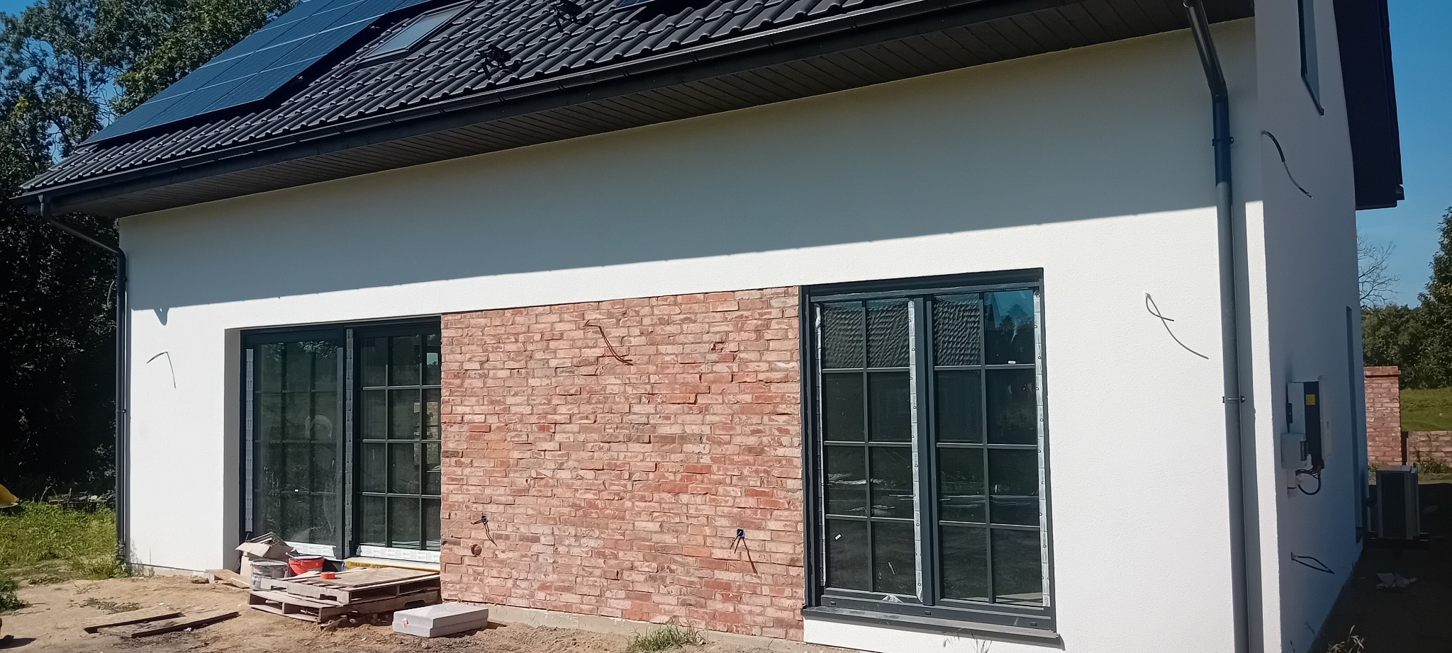 Nowoczesny dom z panelami słonecznymi i fragmentem elewacji wykończonym czerwoną cegłą. Widoczne okna z czarnymi ramami i instalacja elektryczna na zewnątrz.