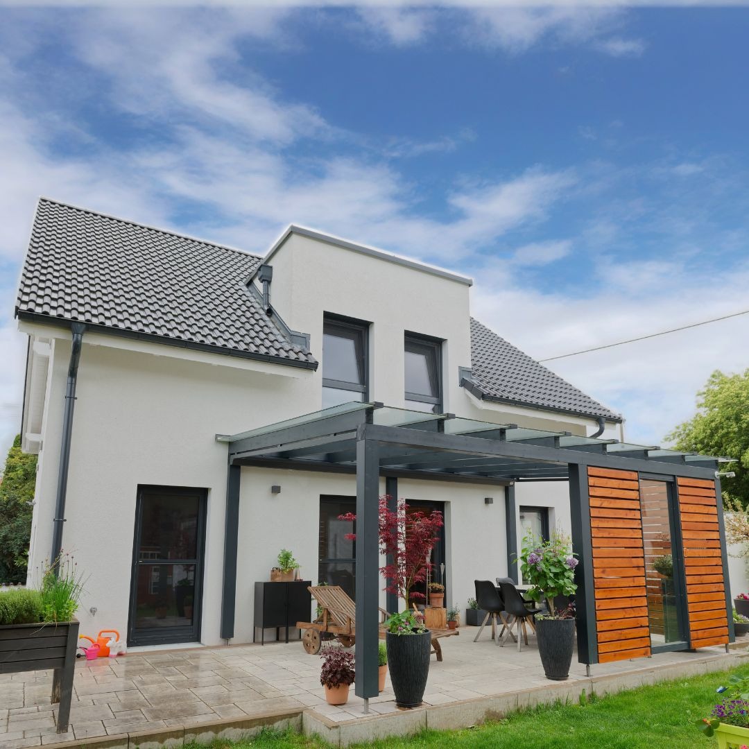 Biały dom z poddaszem użytkowym i szarą dachówką, zadaszony taras z drewnianymi panelami, meble ogrodowe i rośliny w donicach na kostce brukowej.