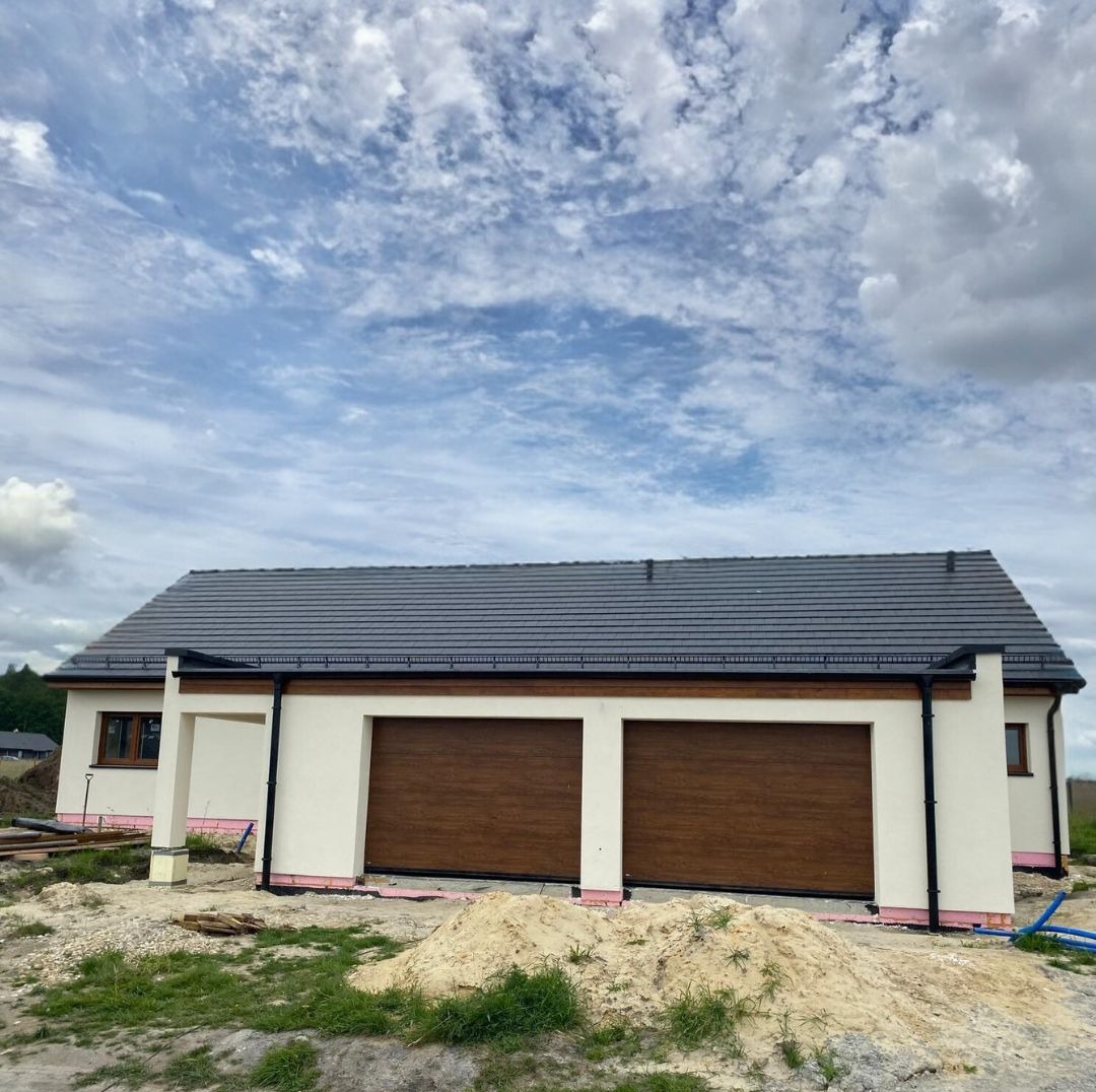 Parterowy dom prefabrykowany z dwustanowiskowym garażem, wykończony z zewnątrz. Widoczne brązowe bramy garażowe i szara dachówka. Na pierwszym planie piasek i trawa.